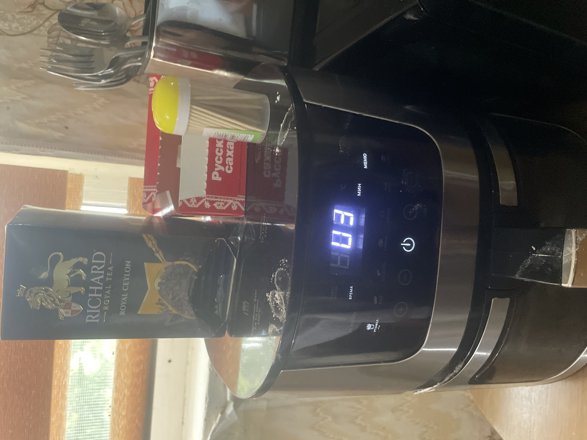 This image shows a close-up of a countertop setting, likely in a kitchen, with several objects visible. A black appliance, possibly an air fryer or a multi-cooker, dominates the right side of the frame, its digital display showing "03". The appliance has several buttons and labels, including "МЕНЮ", "МИН", "ВРЕМЯ", and an icon of a potato, possibly indicating a "fries" or "potato" function. There are some white powdery residues on and around the appliance, suggesting it may have been recently used or is due for cleaning.
To the left of the appliance, a box of "RICHARD ROYAL TEA - ROYAL CEYLON" is prominently displayed. The box features a gold lion emblem and text in English. Further to the left, a red box with Russian text, including "Русск" (Russk) and "саха" (sakha), is partially visible. This box contains what appear to be wooden skewers, capped with a yellow lid. In the upper left corner, a stack of cutlery, including forks and spoons, is held in a metal container.
The overall scene suggests a domestic setting. The lighting appears to be artificial, possibly from an overhead light, casting soft shadows. There are no people visible in the image. The background shows wooden elements, possibly a table or cabinets, and a window with a view of greenery, hinting at an indoor setting during daylight hours. The image is oriented vertically, as if taken with a phone. The visible text includes product names, menu options, and potentially ingredients or instructions in Russian and English.