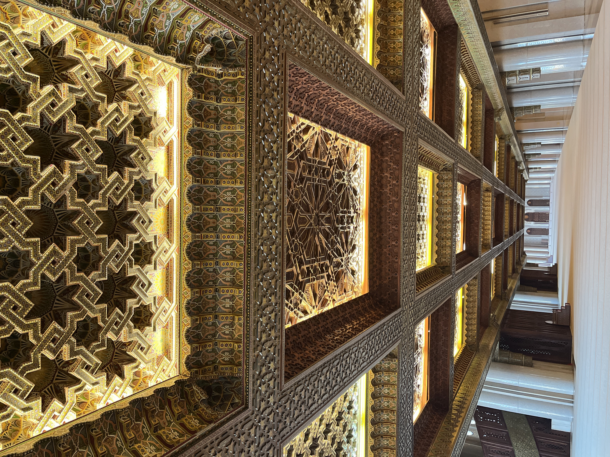 This image showcases a lavish, intricately decorated ceiling, likely found within a grand building in Casablanca, Morocco. The ceiling is adorned with complex geometric patterns, carved from wood and highlighted by a warm, golden light source emanating from behind several panels. These patterns feature interlocking star shapes and arabesques, characteristic of Islamic architectural design.
The ceiling is structured into rectangular and square sections, framed by dark wooden beams that are themselves meticulously carved with smaller, repetitive motifs. The dominant colors are rich browns and golds, with subtle hints of muted greens and reds in painted sections. This ornate detailing covers the majority of the visible space.
Towards the right side of the image, a simpler, light-colored wall or upper building structure provides a stark contrast to the detailed ceiling. The perspective is upward and diagonal, emphasizing the depth and grandeur of the craftsmanship.
There are no people or specific activities depicted in the scene, placing the entire focus on the architectural artistry. The lighting appears to be internal and constant, making the time of day indistinguishable. No visible text or inscriptions are present. The overall impression conveyed is one of awe-inspiring beauty and skilled craftsmanship.