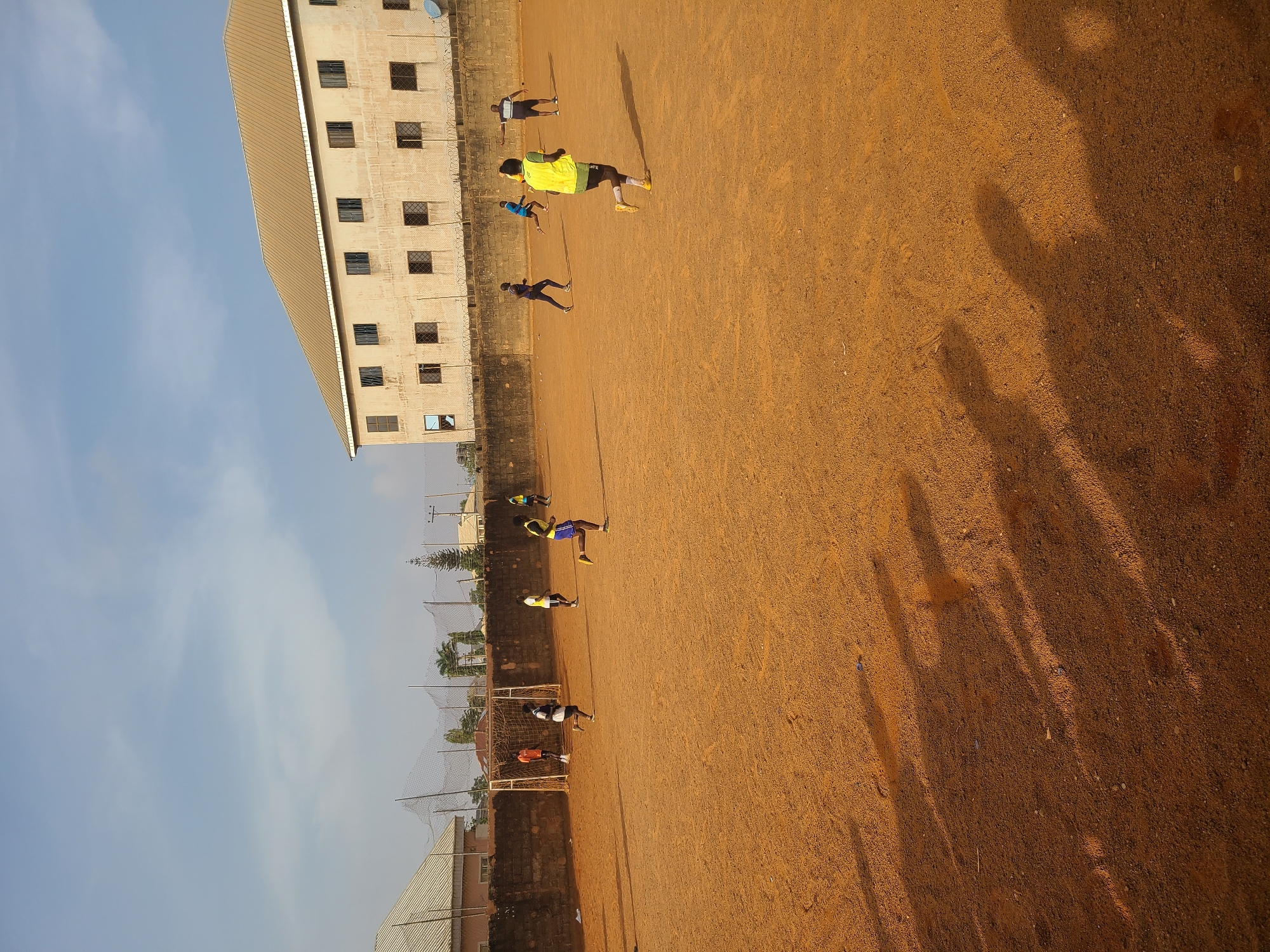 The image depicts an outdoor scene in Barakin Lerawi, Nigeria, showing a group of people engaged in what appears to be a sports activity on a reddish-brown dirt field. The image is oriented vertically, but when viewed upright, the scene features a large, multi-story building with numerous windows on the left side, bordered by a fence, possibly with barbed wire. A satellite dish is visible on the roof of this building.
On the dirt field, several individuals, likely young men or boys, are spread out. One prominent figure in a bright yellow vest and black shorts is near the center, with others in various positions around the field. Their long, distinct shadows stretch across the ground, indicating that the sun is low in the sky, suggesting either early morning or late afternoon. The warm, golden hue of the dirt reinforces this impression.
Towards the far end of the field, a soccer goalpost with a net is visible, confirming the nature of the activity as likely football (soccer) practice or a casual game. Other distant buildings, trees, and tall nets (possibly for ball containment or part of a larger sports complex) can be seen along the perimeter. The sky above is a clear, light blue with some wispy clouds, indicating fair weather. There is no clearly legible text visible on any subjects or objects in the image.