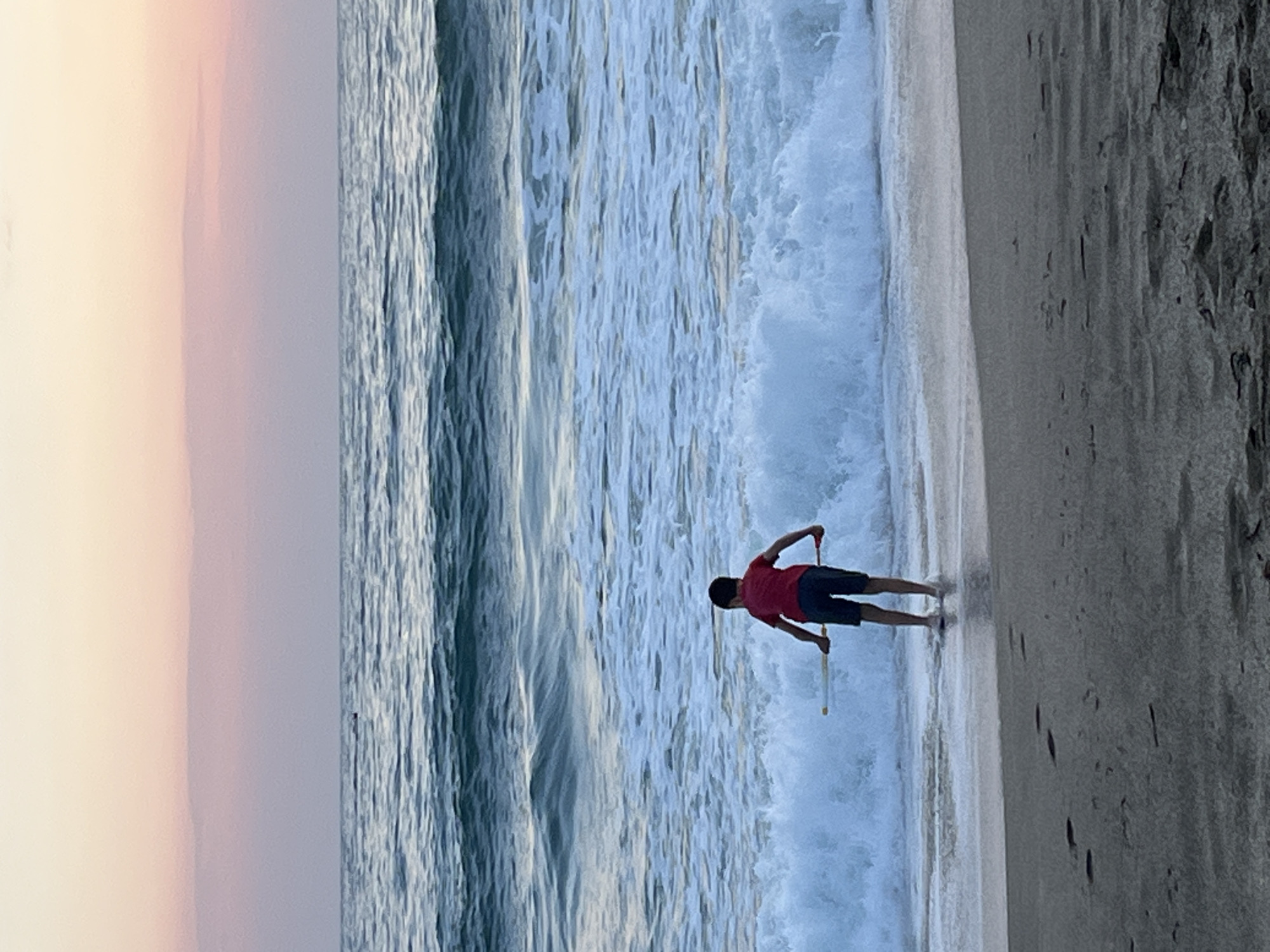 The image depicts a beach scene in Alanya, Turkey. A person wearing a red shirt and dark shorts is standing on the shoreline, near the edge of the water. They are holding what appear to be two thin rods. The water is foamy and active, with waves crashing onto the beach. To the left of the water is a strip of sand, with a pale, faded sky above. To the right is a darker sand area with footprints.