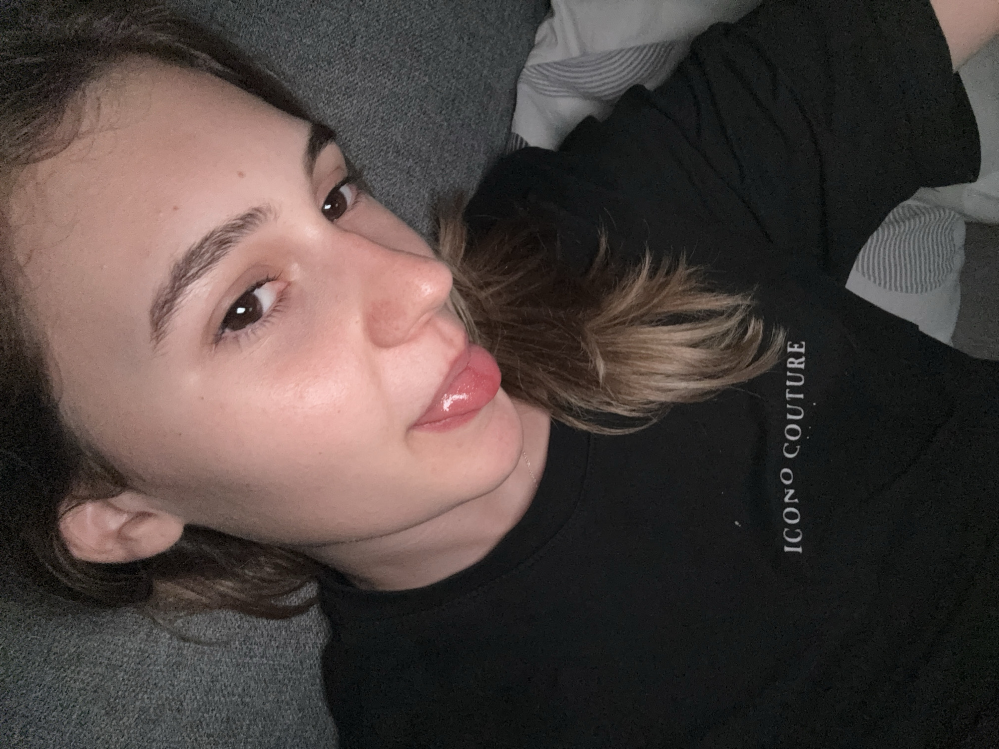 The image shows a close-up of a young woman lying down, with her head resting on a gray surface, possibly a couch or bed. She is wearing a black long-sleeved shirt with the text "ICONA COUTURE" printed in white on the left side of the chest. Her hair is brown and falls over her shoulder. She is looking towards the camera with her tongue slightly sticking out, and her lips appear glossy. The background shows a textured gray surface and a pillow with gray and white stripes. There is no visible indication of the city of Forbach, Germany within the image.