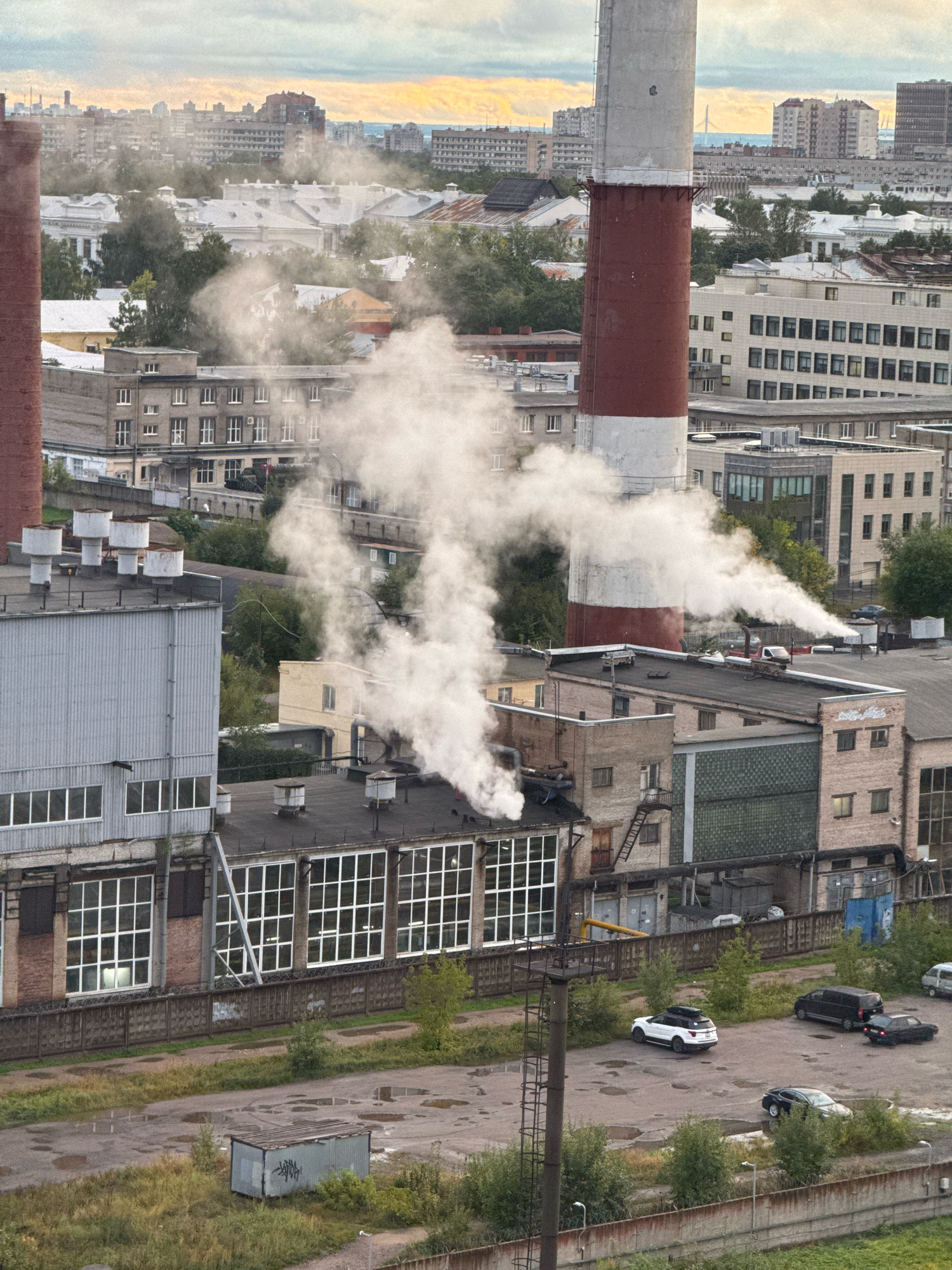 The aerial view captures an industrial area within an urban environment, likely Russia, given the architecture and context. Dominating the center of the frame is a tall, red and white striped smokestack, emitting a substantial plume of white smoke. This smoke billows across the scene, obscuring parts of several buildings and the sky.
Surrounding the smokestack are various industrial structures with flat roofs and rows of windows. Some buildings are made of brick, while others have metallic siding. Several smaller vents or chimneys are visible on the rooftops, along with air conditioning units. A network of pipes and elevated walkways connects some of the structures.
In the foreground, a paved area, possibly a parking lot, shows signs of disrepair with puddles of water and patches of overgrown grass. Several cars are parked here, including a white SUV, a black minivan, and a dark sedan. A small, dilapidated metal shed with graffiti markings sits on the grass to the left. A chain-link fence with barbed wire runs along the edge of the industrial area.
In the background, a dense cityscape with residential and commercial buildings stretches towards the horizon under a cloudy sky, with hints of yellow and orange suggesting a sunset or sunrise. The overall impression is one of active industrial production within a developed urban setting. There are no people visible in the image.