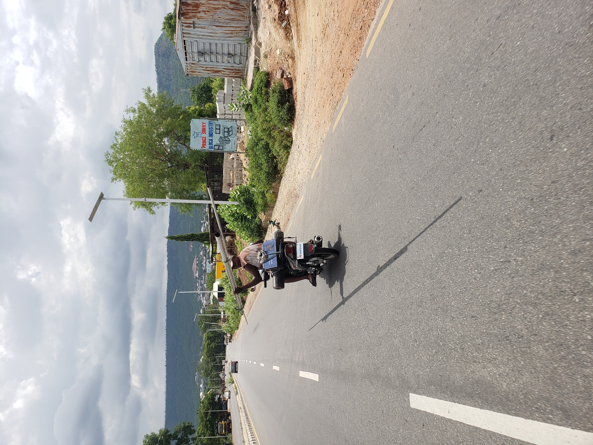 The image, correctly oriented, displays a daytime scene in Adankolo, Nigeria, featuring a person on a motorcycle on a paved road. A man, wearing a light top and dark bottoms, is seen either riding or stopped on a dark-colored motorcycle, actively transporting several long, metallic poles or pipes. These poles are strapped to the side of the motorcycle and also held by the rider, extending forward. The motorcycle's license plate is visible and reads "W XIAOXUE".
The asphalt road has clear white and yellow markings. A long shadow cast by the motorcycle and its rider stretches across the road, indicating it is daytime, likely late morning or early afternoon. To the right of the road, there is a dirt shoulder with green bushes and a small tree. Further back, a rustic, corrugated metal shed or building stands, and stacks of concrete blocks are visible. A white sign with blue lettering reads "PRINCE SOMAY BLOCK INDUSTRY" and includes a phone number, "07033327660", along with an illustration of concrete blocks. A street light pole with a solar panel attachment is also present.
The background is dominated by lush green mountains or hills beneath a heavily clouded sky. Other vehicles are faintly discernible in the distance along the road. The scene captures an everyday moment of transportation and local industry.