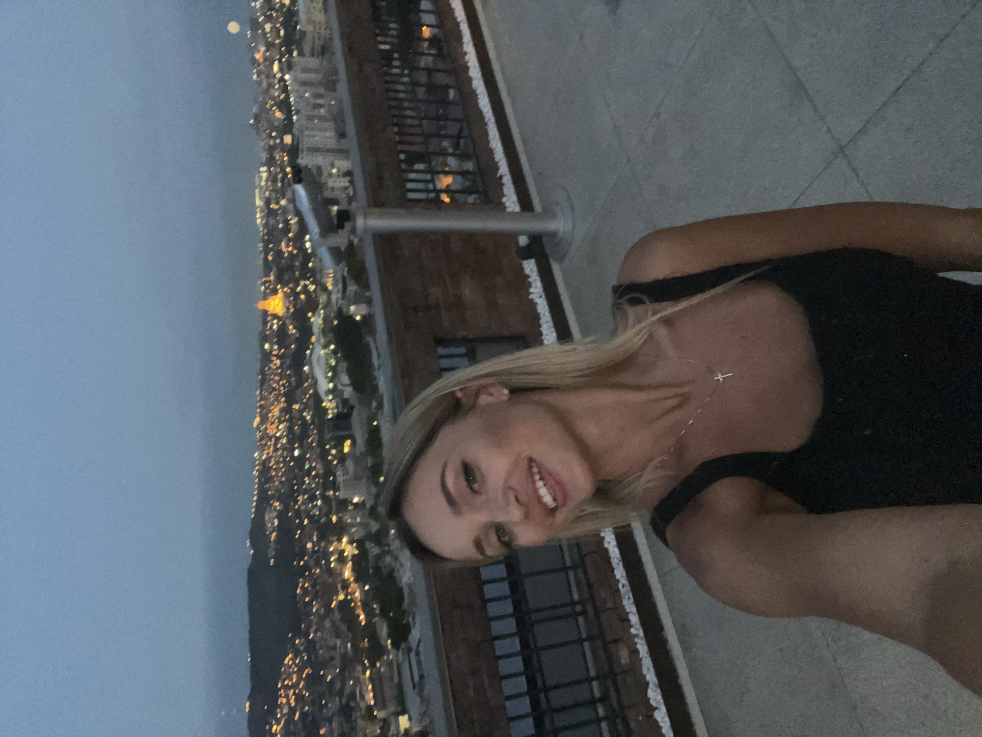 A blonde woman in a black tank top smiles at the camera with her tongue sticking out slightly. She is wearing a silver necklace with a cross pendant. She is standing on a rooftop terrace overlooking a city at dusk. The city lights are on, and the sky is a deep blue. In the distance, there is a bright orange-domed building and the moon is visible in the sky. A telescope is mounted on the terrace railing. The terrace has a stone floor and a brick wall with a metal railing.