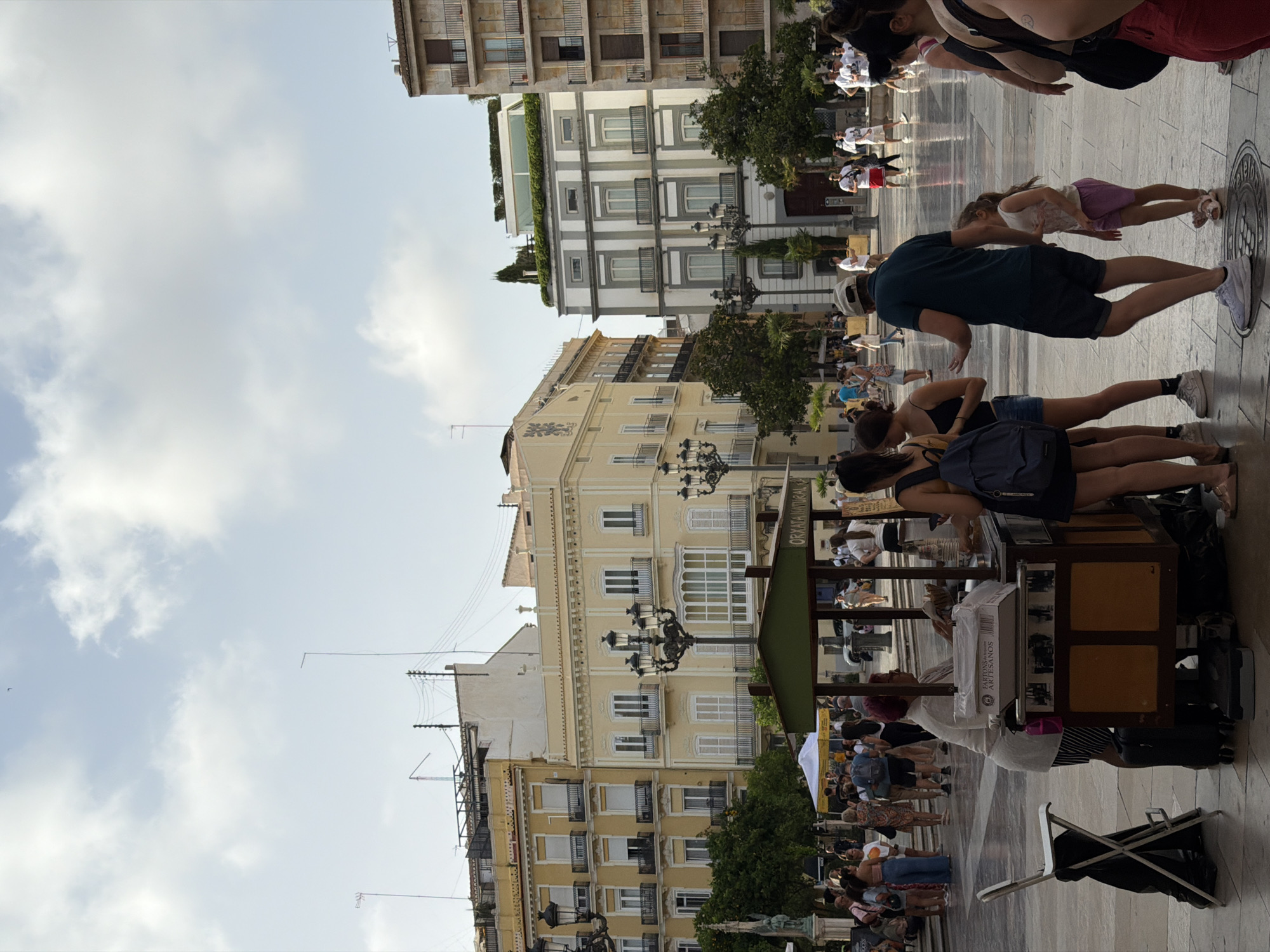 In a public square, likely in a European city based on the architecture, people are gathered around a food cart. The cart, which appears to sell "ORXATA NATURAL," has a green canopy and is attended by a woman with red hair. Several people are interacting with the cart, with one woman in the foreground on the right looking towards it. In the background, other individuals are walking and standing, and buildings with balconies and windows line the square. The sky is partly cloudy, and the ground is paved.