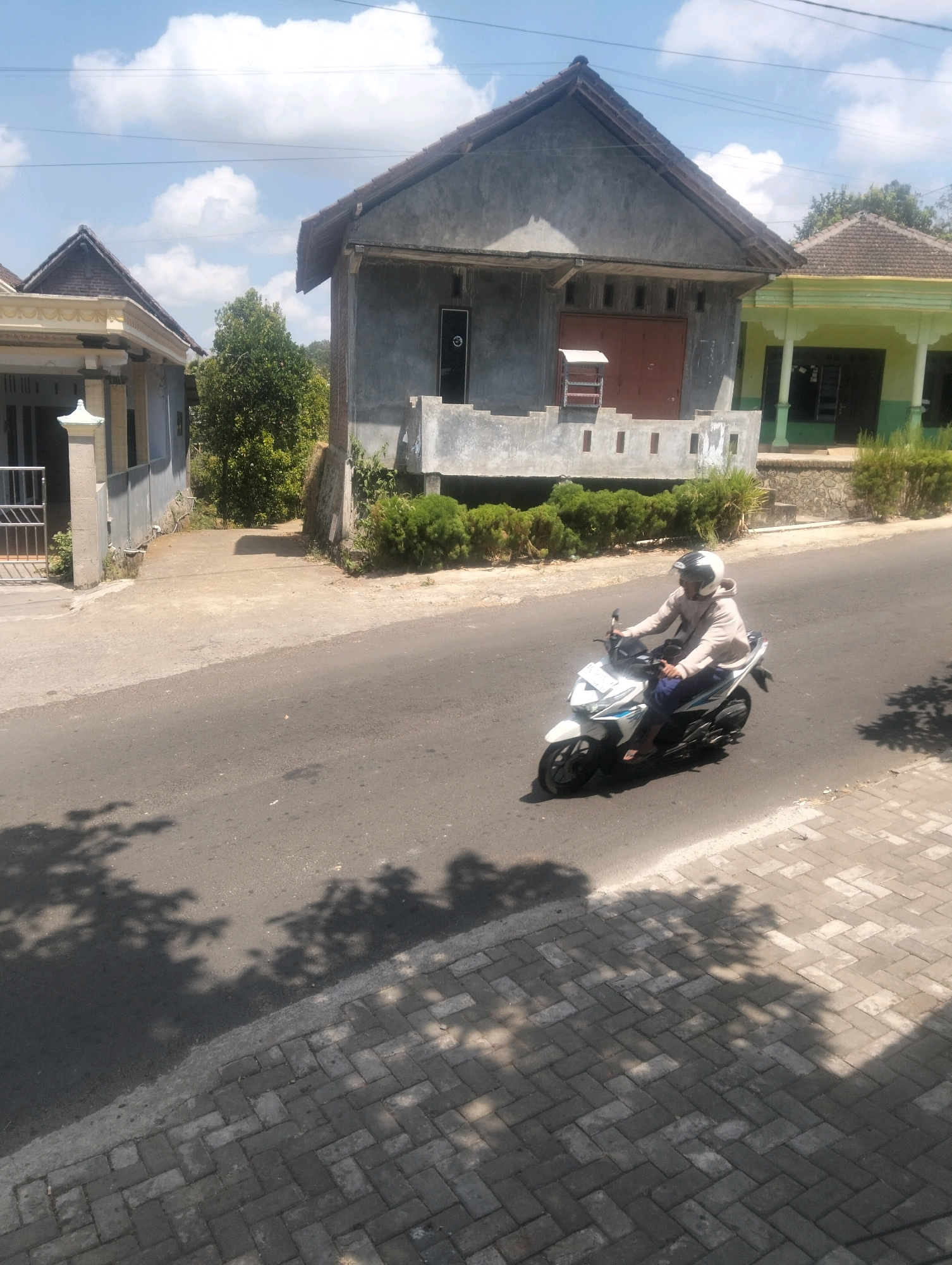 In Kanyoran, Indonesia, a person wearing a light-colored hooded sweatshirt and a white helmet rides a white scooter on an asphalt road. The scooter has blue and black accents. The rider is moving from left to right in the frame. The road is bordered by a paved walkway made of grey rectangular bricks on the right side of the image. Tree shadows are cast across the road and walkway. In the background, there are several houses with gray and light green exteriors and tiled roofs. A dense green hedge runs along the front of the houses. The sky is blue with scattered white clouds.