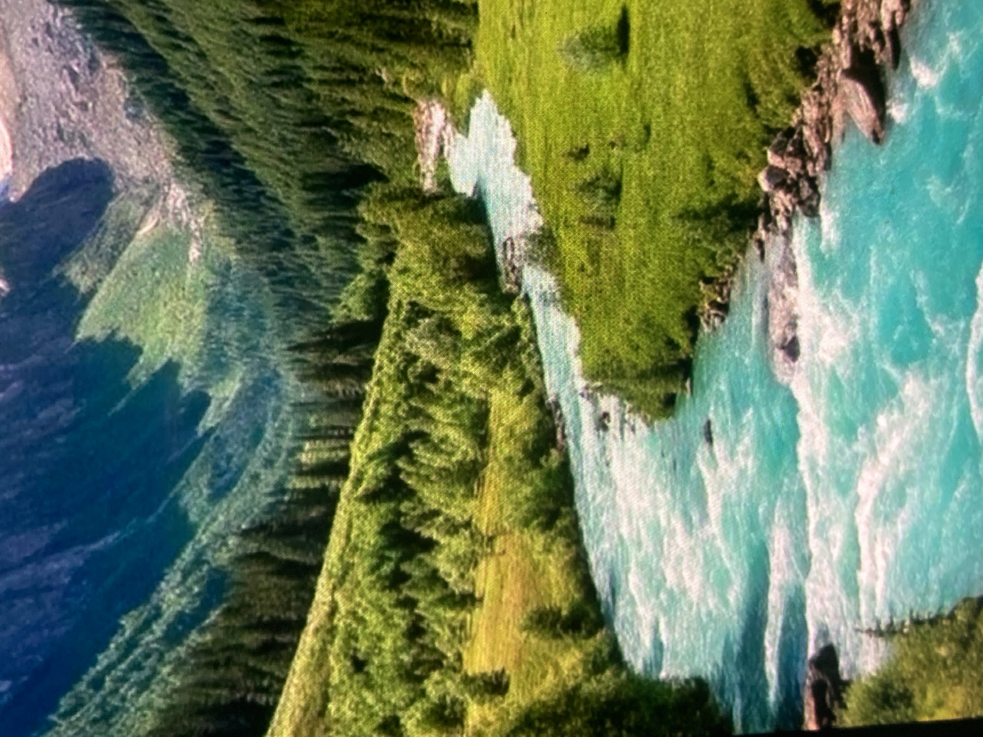 The image shows a vibrant, turquoise river winding through a lush, green valley. The river has numerous rapids and white water, suggesting a fast flow. The valley slopes upwards on either side, covered in dense green forests and meadows dotted with scattered trees. In the distance, rugged, rocky mountains rise, their peaks partly shrouded in shadow and light. The scene is a natural landscape, characteristic of alpine or mountainous regions. There are no people or man-made structures visible. The location is specified as Ustyvytsia, Ukraine, implying this natural scene is found within or near this Ukrainian settlement.