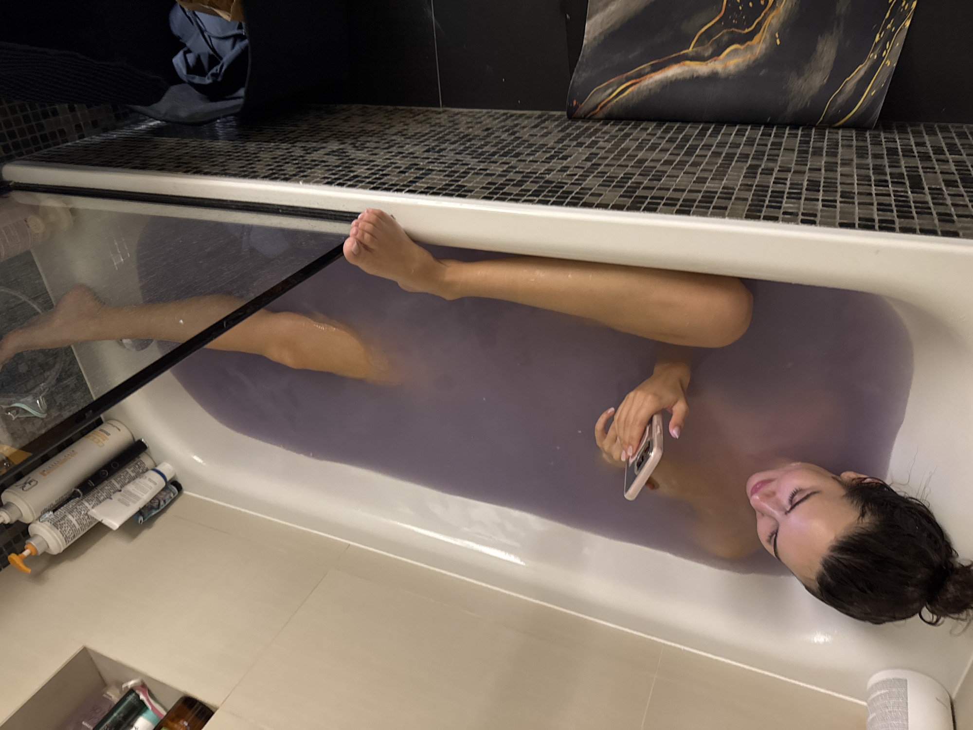 A woman is depicted relaxing in a bathtub filled with purplish water. She is lying on her back, with her head and most of her torso submerged, while her legs are stretched out along the tub. Her dark hair is pulled back into a bun, and she has a calm expression, looking down at a light-colored smartphone with a clear or light-colored case that she holds in her right hand. Her fingernails are painted a light color.
The bathtub is white, and several bottles and tubes, including some with pump tops, are lined up on the edge of the tub near her feet. A black cable, possibly a phone charger or headphone cable, runs from near her left arm towards the left side of the tub.
The bathroom features a wall with small, dark mosaic tiles behind the tub. On the tiled ledge above the tub, a dark pillow with abstract gold and orange patterns rests. Another piece of dark fabric, possibly clothing or a towel, is visible on the left side of the ledge. The floor tiles are large and light-colored. A white gridded object, possibly a vent, is partially visible in the bottom right corner of the frame. The lighting appears artificial, suggesting an indoor setting, likely in the evening or night. No specific text is clearly legible on any of the objects.