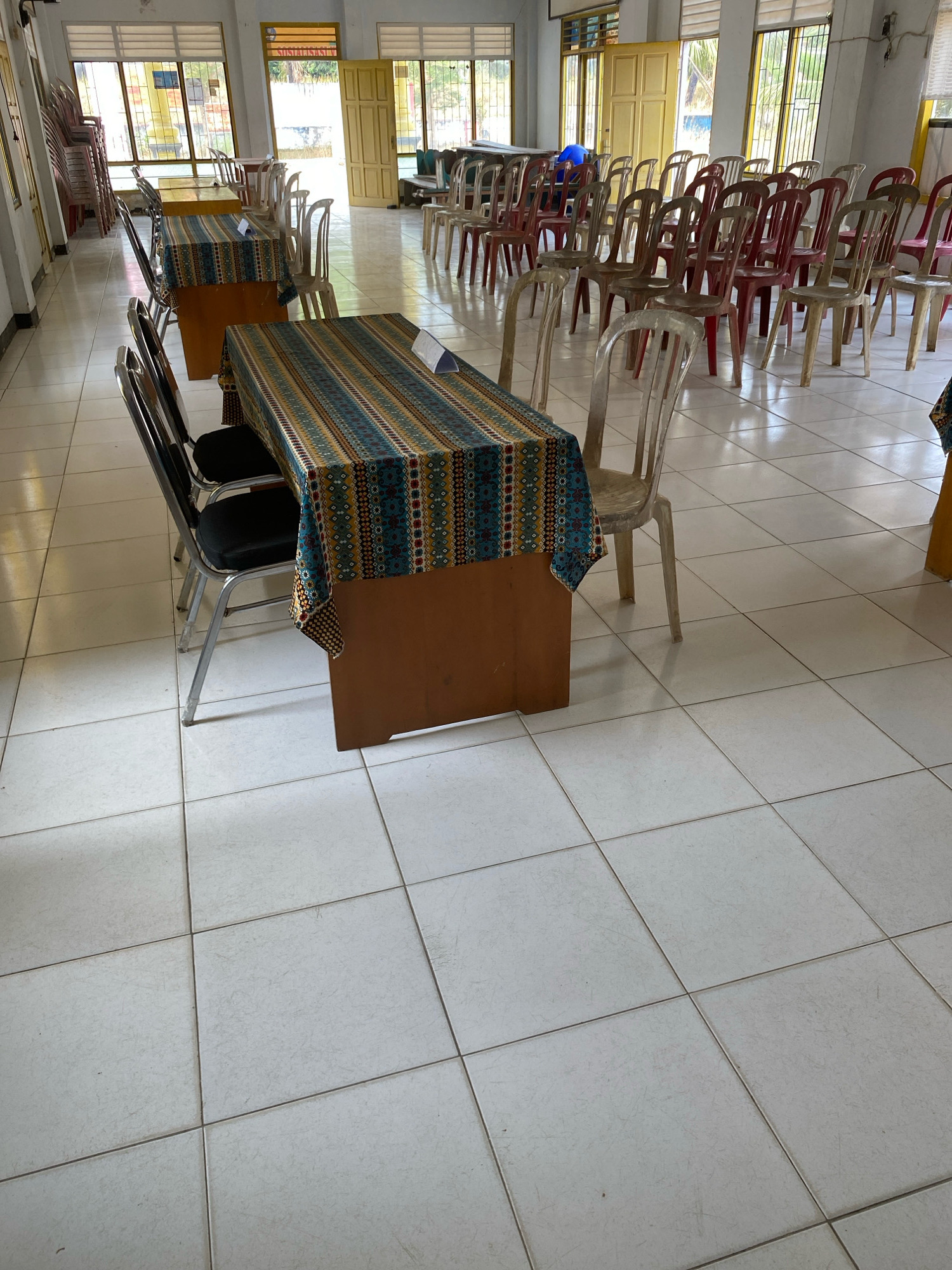 The image depicts a room with tiled floors and windows. There are tables covered with patterned cloths and chairs set up around them. In the background, stacked chairs and open doors leading outside are visible. The scene appears to be set in a communal or event space. The specific location within Mojo Pahit, Indonesia, cannot be determined from the image alone.