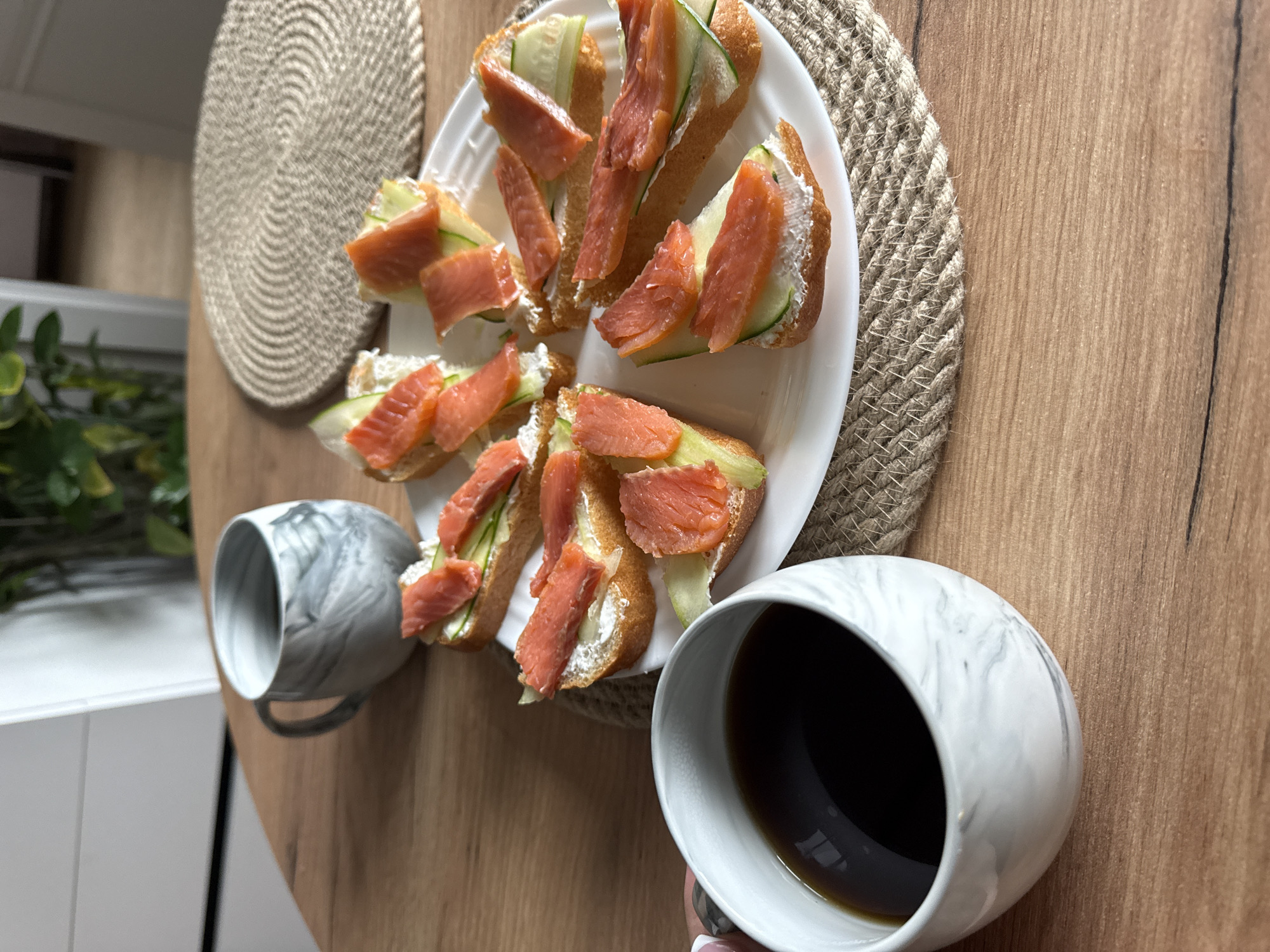The scene presents a casual food setting. A white plate occupies the center of the view, carrying several open-faced sandwiches, seemingly composed of bread slices, cream cheese or similar spread, cucumber, and salmon. The plate sits on a woven round mat, which in turn rests on a wooden table with a light brown finish.
Two marble-patterned mugs are placed on the table, one filled with a dark liquid, presumably coffee, and the other empty.
In the background, visible on the left side, there's a hint of green foliage, possibly from a potted plant, and a white sideboard or cabinet.
Given the information in the filename, this scene is likely located in Новая Разводная, Russia. No people or specific actions are evident.