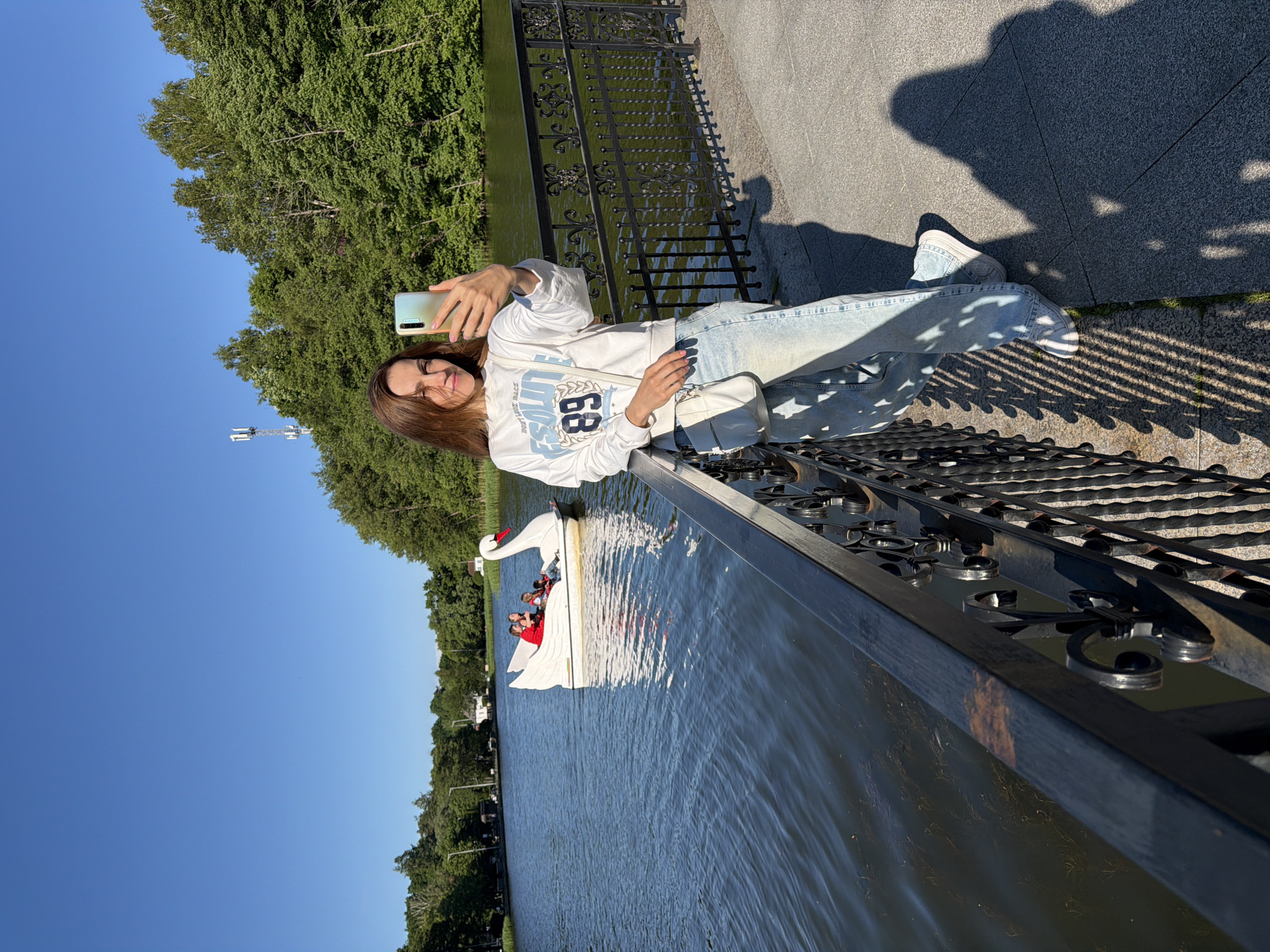 A young woman with long brown hair stands on a paved walkway next to a black metal railing. She is taking a selfie with a smartphone held in her right hand. She is wearing a white sweatshirt with blue lettering and light-wash jeans. A white crossbody bag is slung over her left shoulder.
Behind her, a calm body of water reflects the clear blue sky. On the water, a white swan-shaped paddleboat is carrying at least three people. The paddleboat is positioned in the mid-ground.
The background is a dense line of green trees. A cell tower is visible on the left side of the image, rising above the trees. The walkway is made of grey stone pavers, and a shadow from the railing is cast across it. The scene appears to be a park or recreational area in Svetlogorsk, Russia, likely near a lake or river, given the presence of the paddleboat and the natural landscape. The lighting suggests it is daytime, possibly late afternoon due to the long shadows.