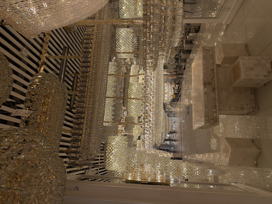The image depicts an opulent indoor showroom, likely a luxury lighting or chandelier store, in Dubai, United Arab Emirates. The space is filled with hundreds of sparkling crystal chandeliers of varying shapes and sizes, suspended from the ceiling and extending far into the background, creating a dazzling, immersive display.
The chandeliers are intricately crafted from numerous individual crystal pieces, forming cylindrical, rectangular, and more complex multi-tiered designs that reflect and refract light intensely. The ceiling above them features a distinctive black and white striped pattern, providing a bold, contrasting backdrop to the shimmering fixtures. The floor below is highly polished and reflective, possibly made of marble or polished tiles, mirroring the intricate light patterns from the chandeliers.
On the right side of the image, several substantial, block-like display stands or pedestals, constructed from light-colored marble or stone, are visible. Some of these have textured or inlaid surfaces. In the distance, at the end of the long aisle of chandeliers, a few indistinct, blurry figures can be discerned, suggesting customers or staff exploring the vast collection. The scene conveys an atmosphere of immense luxury and grandeur. No legible text is visible on any tags or signs.