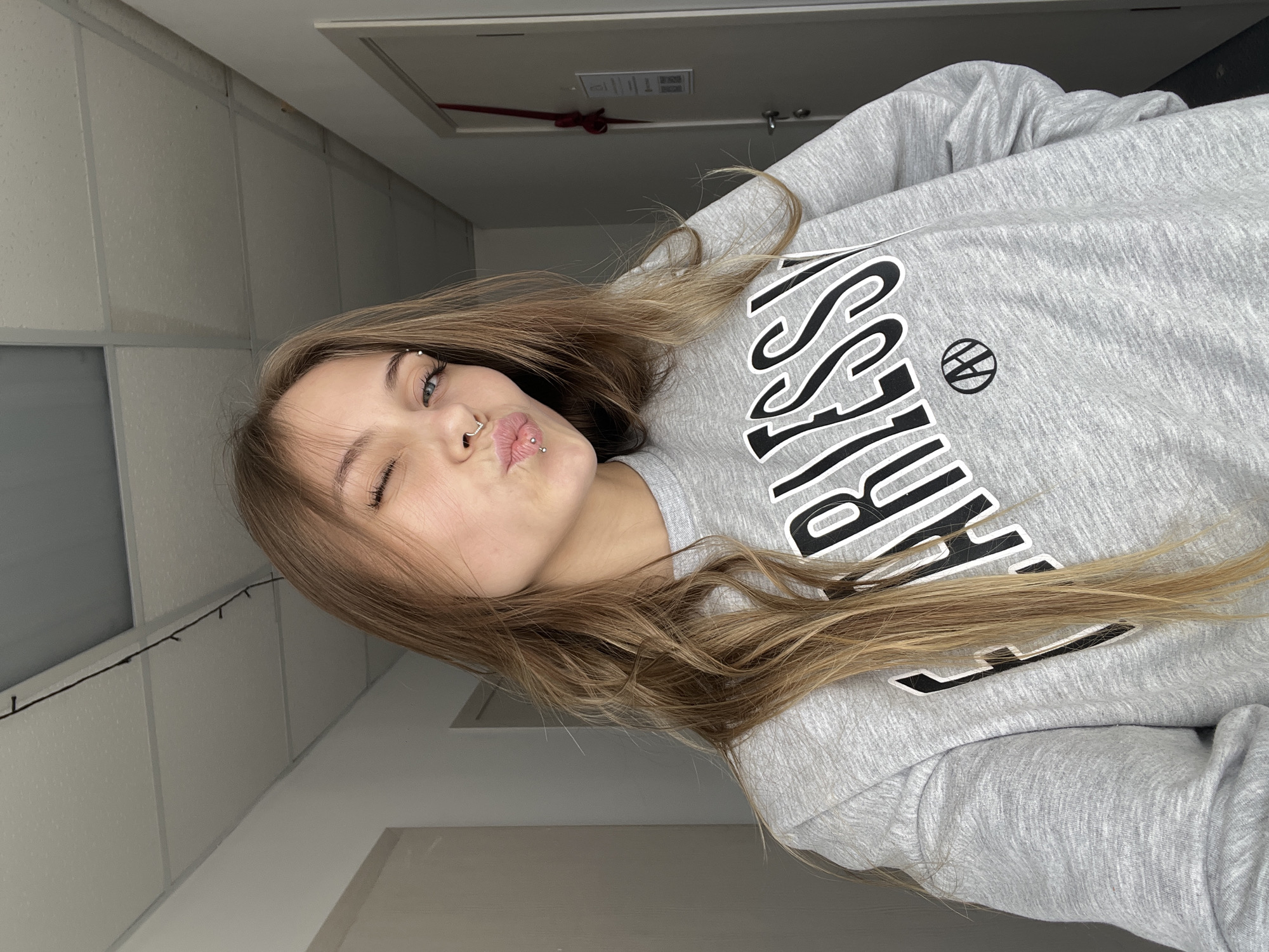 A young woman with light brown hair, blue eyes, and multiple facial piercings, including an eyebrow piercing, nose ring, and lip piercing, is shown in an indoor setting. She is wearing a grey sweatshirt with the word "FEARLESS" printed on it in large black letters. The background appears to be a hallway or room with white walls and a suspended ceiling with rectangular panels. There is no verifiable information to indicate the location is in the city of Фоки, Russia.