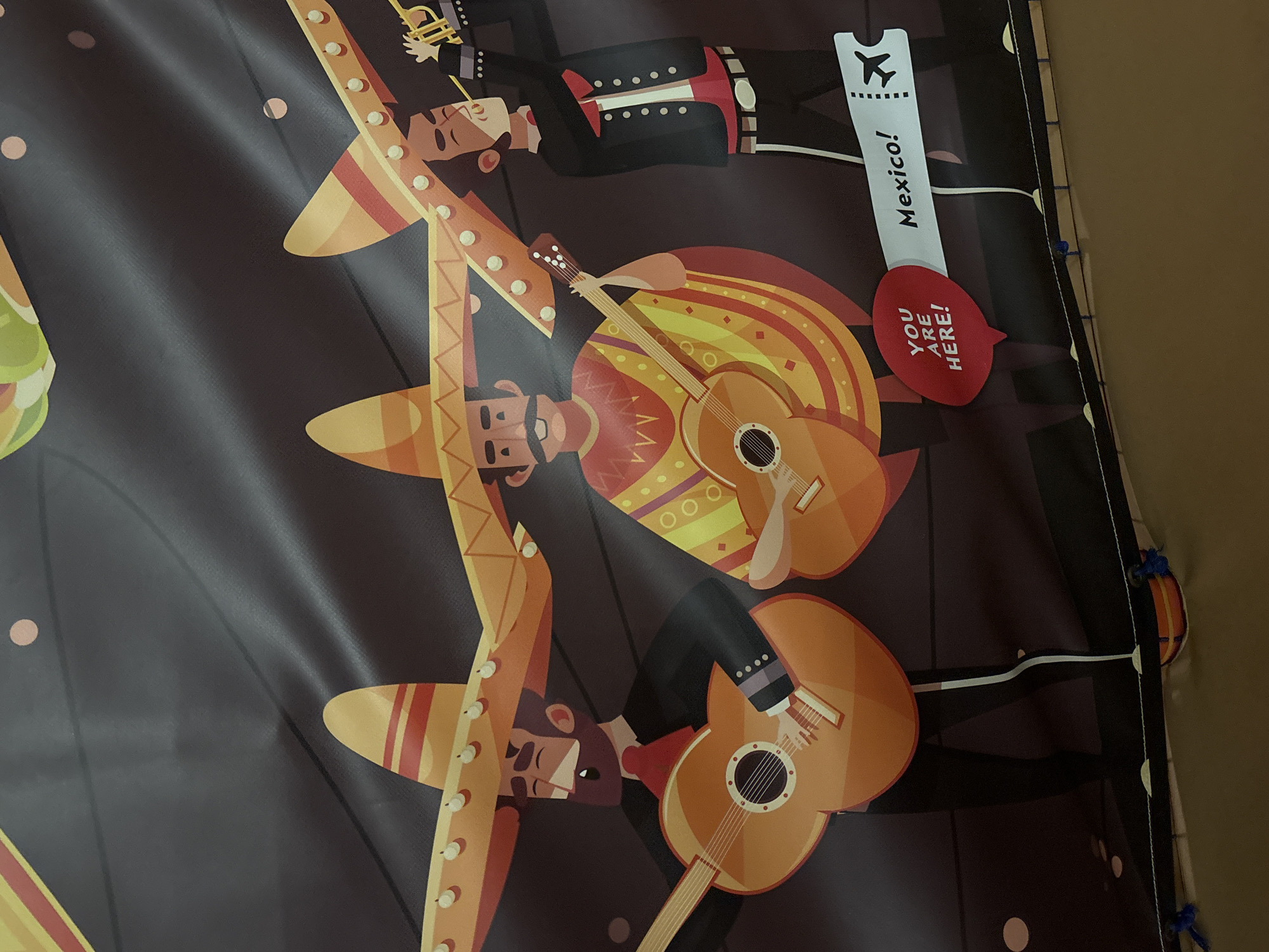 The image shows a close-up, slightly angled shot of a banner or backdrop featuring three cartoon mariachi musicians against a dark, textured background. The musicians are depicted in a stylized, flat illustration style.
From left to right:
The first mariachi, on the far left, wears a large sombrero, a red tie, and a black jacket. He is playing an orange acoustic guitar.
The second mariachi is in the center, also wearing a sombrero and a colorful serape. He is also playing an orange acoustic guitar.
The third mariachi, on the far right, wears a sombrero and a black suit with red accents. He is playing a trumpet.
In the lower right portion of the image, there is a stylized ticket stub that reads "Mexico!" with an airplane graphic. To the left of the ticket is a red speech bubble that says "YOU ARE HERE!".
The background appears to be a dark fabric that is wrinkled and creased, suggesting it is a banner or a similar display. There are some circular, light-colored dots scattered on the background, possibly representing confetti or stars. The bottom of the image shows a textured, brown surface, likely a floor or table, and the edge of the banner is visible with some blue ties attached.
This banner is not located in Dubai, United Arab Emirates. Based on the imagery, it is likely a decorative item for a Mexican-themed event or establishment.