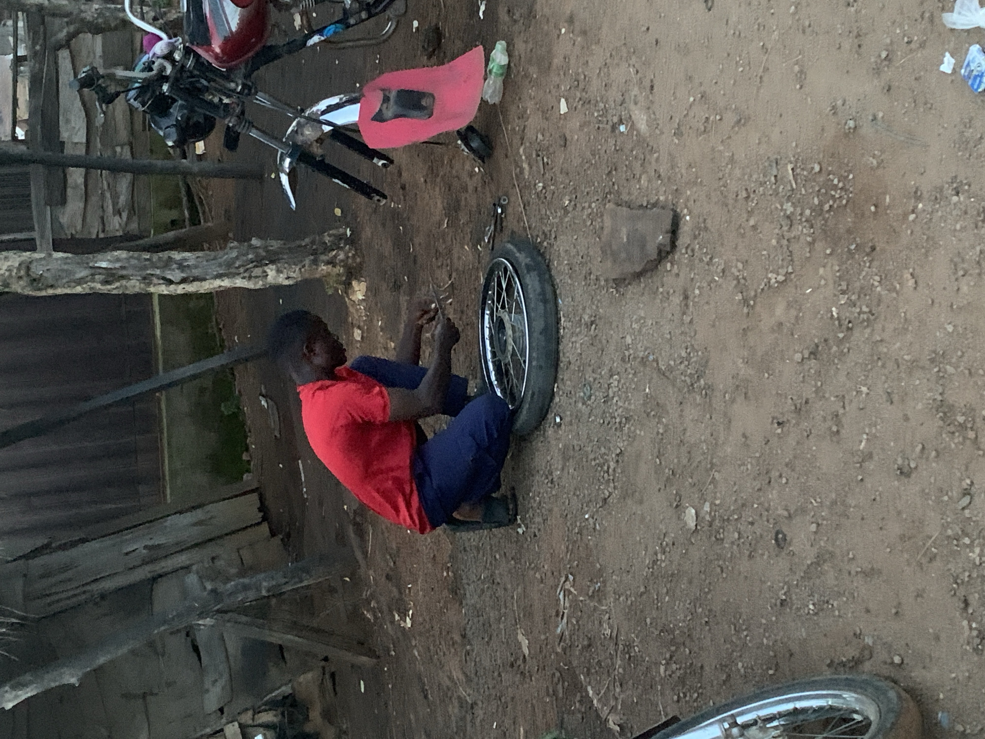 In Gbeleko, Nigeria, an outdoor scene depicts a male individual performing mechanical work. The subject, an adult male with dark skin, wears a red short-sleeved shirt, dark blue trousers, and dark sandals. He is kneeling on a reddish-brown earth and gravel surface, focused on a spoked motorcycle wheel lying flat before him. He holds small metal tools, consistent with tire manipulation or removal, near the wheel's rim.
To the right, the front section of a red and black motorcycle is partially visible; it lacks its front wheel. The motorcycle's handlebars, front forks, and a portion of its fuel tank are discernible. A red fabric object is draped over the motorcycle's front forks. A light green plastic bottle lies on the ground nearby.
The background on the left features weathered wooden structures, constructed from planks and beams. On the right, a tree trunk with textured bark is visible, with additional indistinct structures behind it. The immediate foreground on the lower left shows a portion of another spoked wheel with a tire. The ground is unpaved, scattered with gravel, small stones, and minor organic debris. The scene is illuminated by ambient daylight.