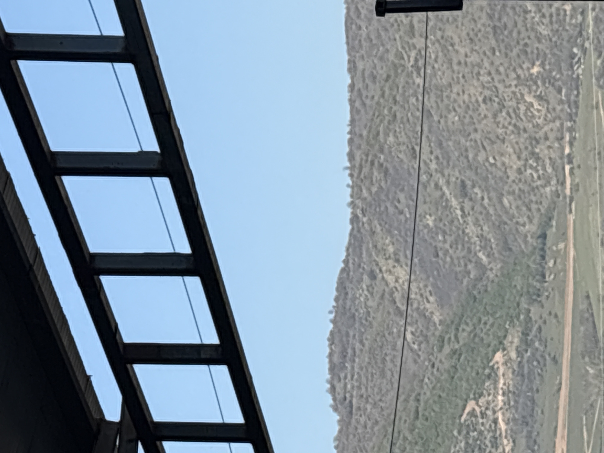 The image shows a view from under a partially constructed or derelict metal structure, possibly a building or overhead cover, towards a forested mountain slope in the background. The sky is visible through the rectangular openings of the metal frame. A power line or cable runs across the view towards the mountain. The location is potentially Struma, Bulgaria.