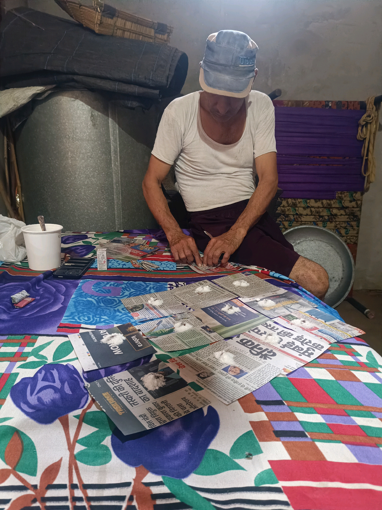 An older man wearing a white tank top and maroon shorts sits on a bed covered with a colorful, patterned sheet. He has a blue cap on his head and is looking down at newspapers spread out on the bed in front of him. On the newspapers, there are small piles of white powder, and the man appears to be working with them. To his left, there is a white cup with a spoon in it, a small black device, and some small packets. In the background, there are stacked items including what looks like rolled-up dark fabric, a woven basket, and purple fabric. A large metal basin is visible to the right of the man. The room appears to be dimly lit.