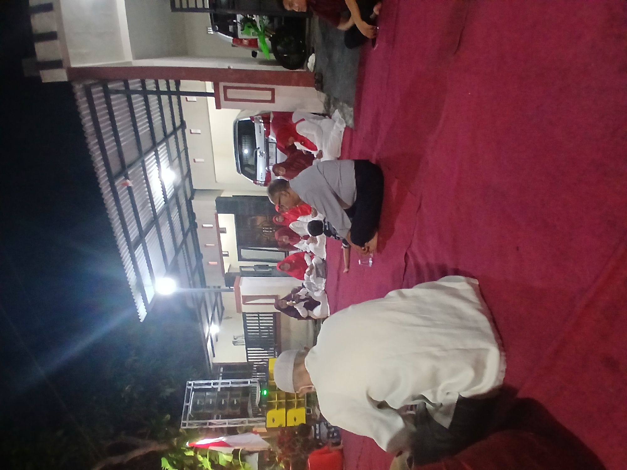 A group of people are gathered on a red carpeted area at night, with a corrugated metal roof structure overhead. The setting appears to be an outdoor gathering, possibly a religious or cultural event, in Tenggarang, Indonesia. In the foreground, a man in a white shirt and cap is seated, with his back to the camera. To his left, a group of women wearing red and white hijabs are seated on the carpet. Behind them, a silver car is parked. To the far left, there are stacks of yellow audio equipment, and a red and white flag is visible. A person in a maroon shirt is seated on the right side of the frame. The overall atmosphere is that of a communal gathering at dusk or night.