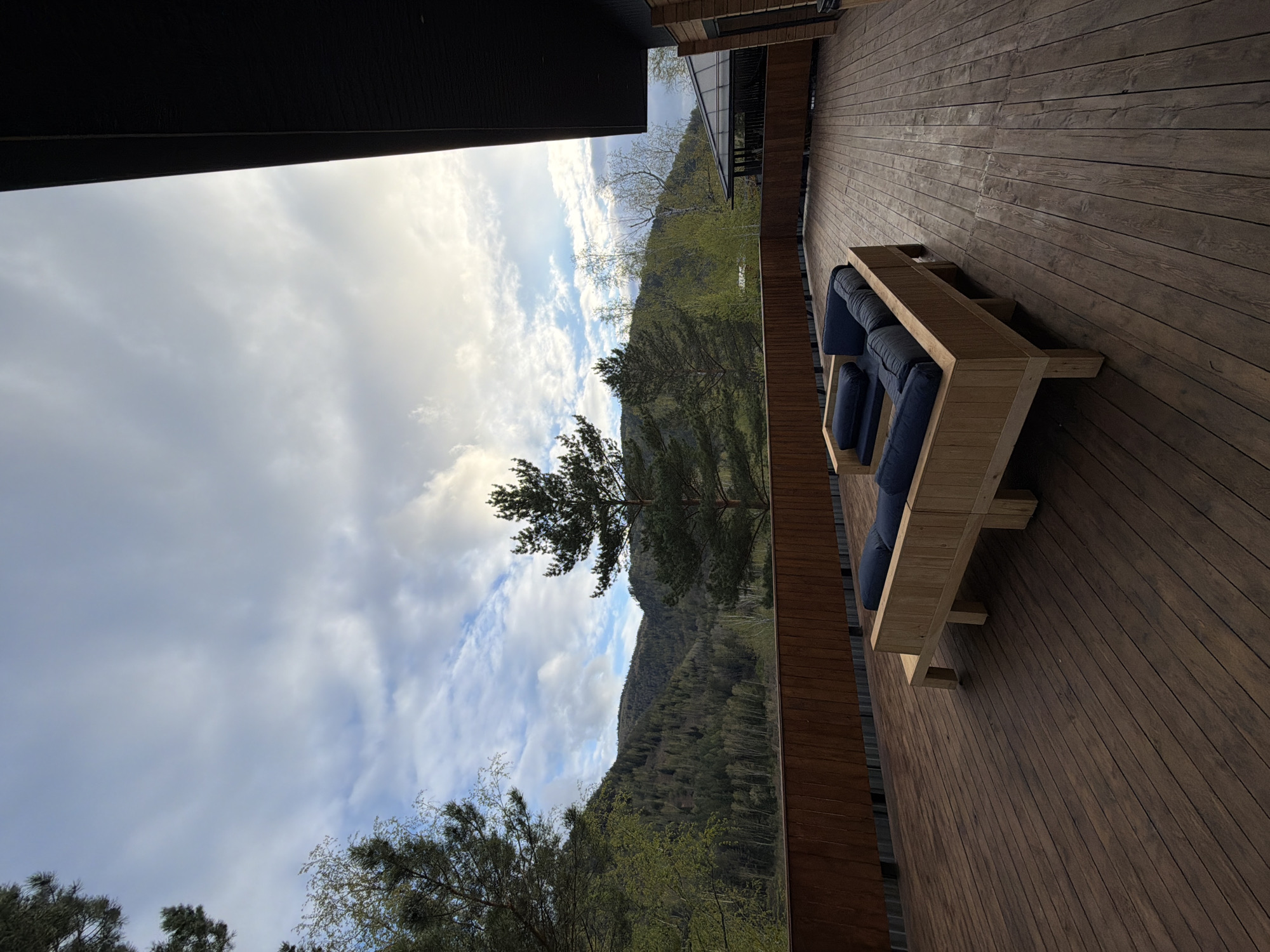The image shows a wooden deck with a low, wooden lounge chair with blue cushions. The deck overlooks a mountainous landscape with trees and a cloudy sky. The scene is framed by architectural elements. It appears to be located in the city of Манский, Russia.