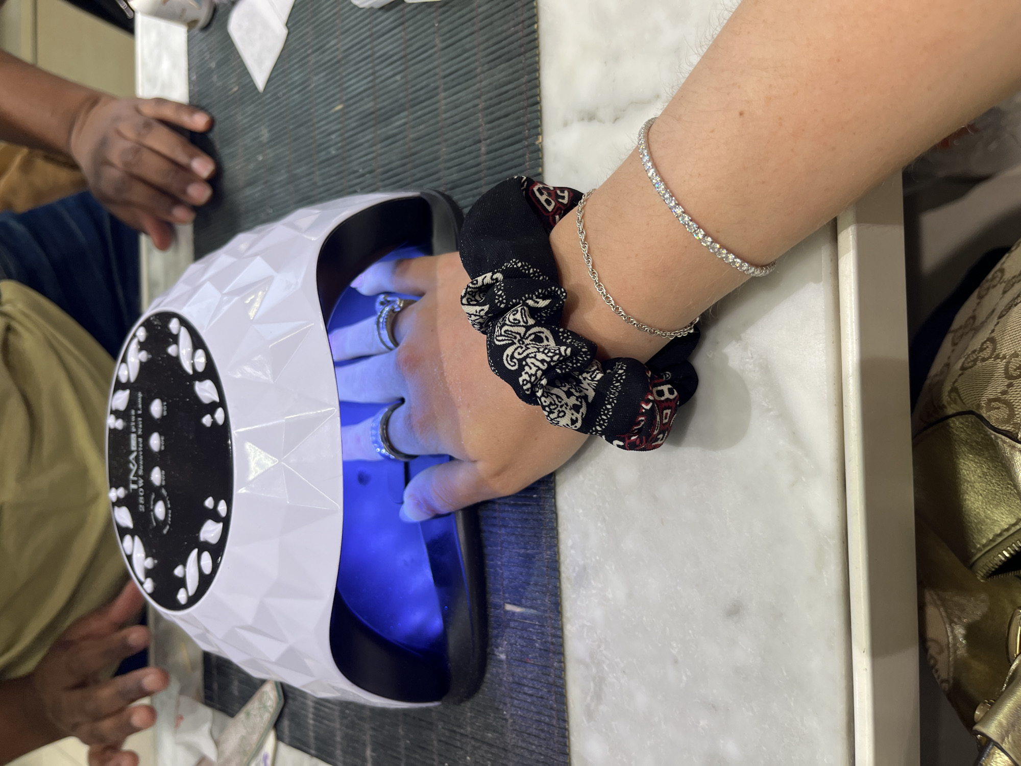 The image shows a person's hand placed inside a white TINA plus 280W Sunuvated Nail Lamp. The person is wearing rings on several fingers and a scrunchie and two bracelets on their wrist. Another person's hand is visible to the left of the lamp. The location is likely a nail salon, based on the presence of the nail lamp and the presumed actions. The image does not provide any information to identify if the location is in Johannesburg, South Africa, or anywhere else.