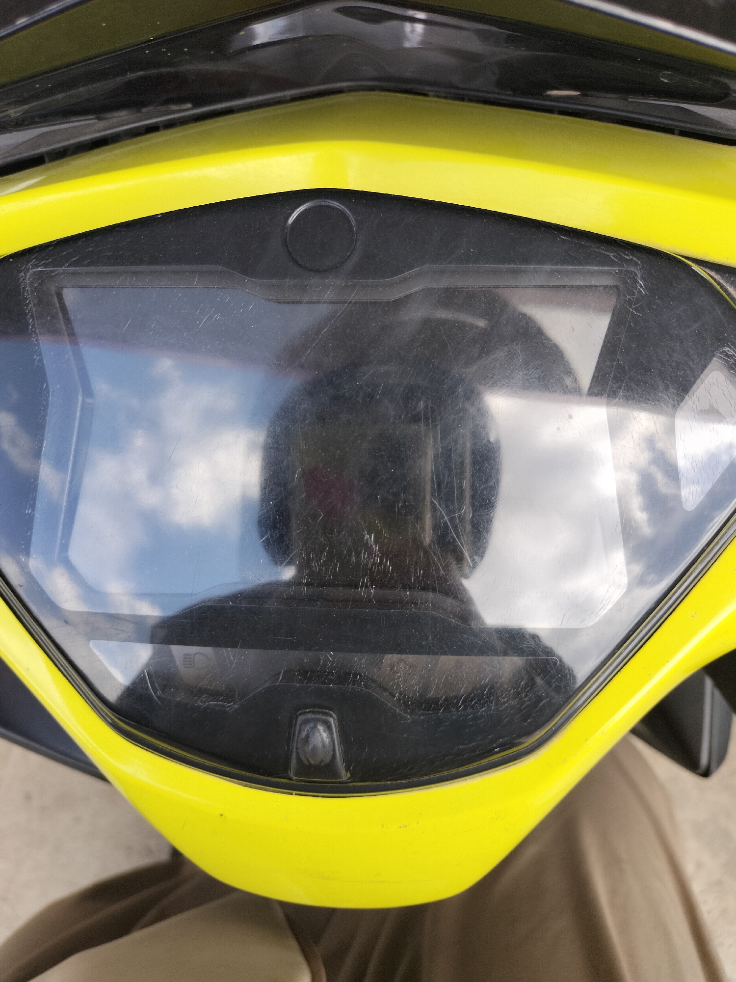 The image is a close-up, top-down view of a yellow motorcycle dashboard with a reflective screen. The screen displays a reflection of the sky with clouds and the rider's helmet. The dashboard is made of black plastic and has some visible scratches on the screen. Below the dashboard, the rider's legs are visible, covered in light brown fabric. The scene appears to be outdoors, possibly in a city setting in Angsana, Indonesia, given the context provided.