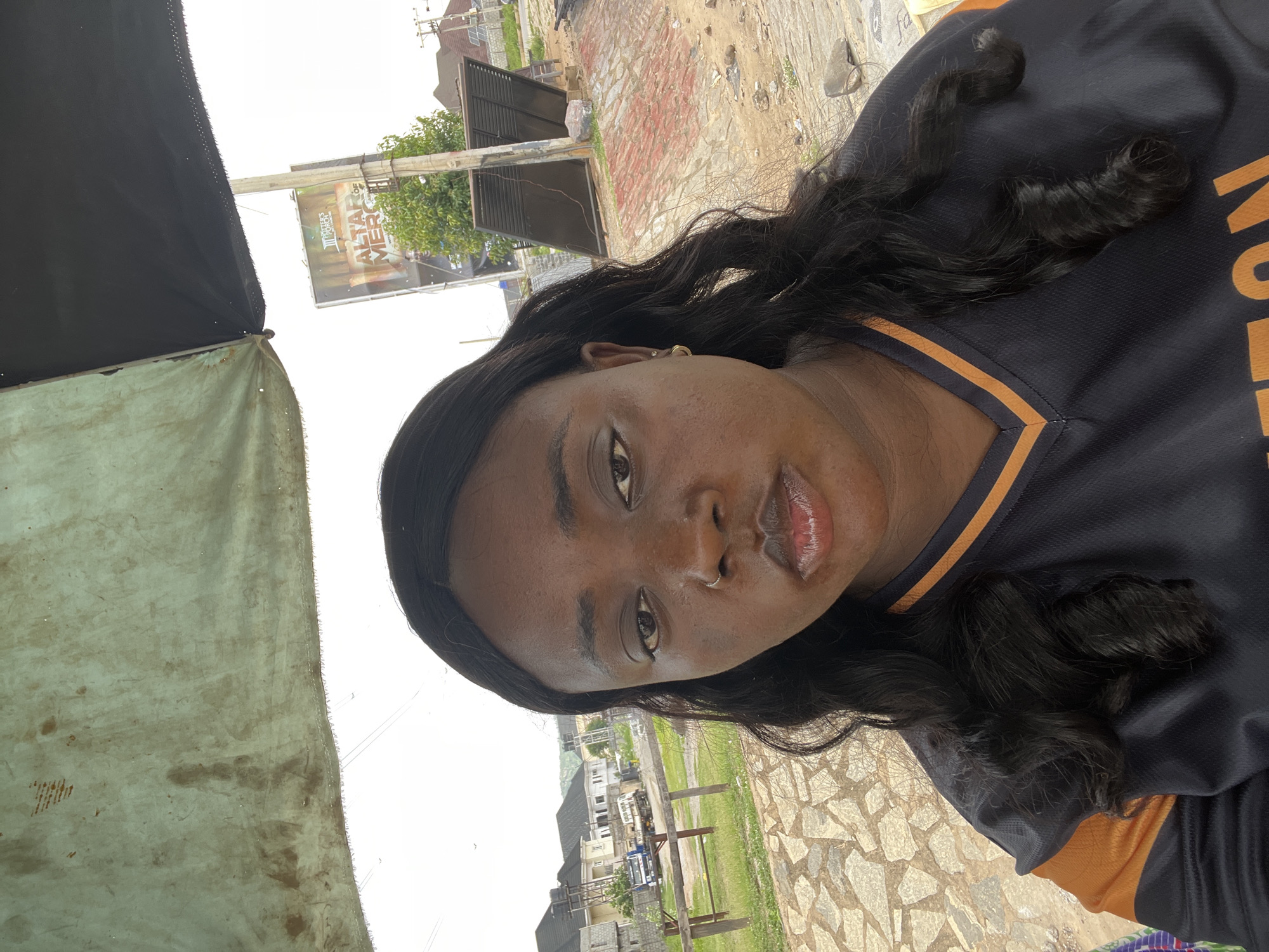 A medium-close-up of a dark-skinned woman wearing a black and orange shirt. She has long, dark hair and is wearing earrings. The background includes a worn-looking awning on the left and a stone-paved area with grass and buildings further back. A billboard is visible in the distance. The location is likely in Abuja, Nigeria, based on the environment and architectural style.
