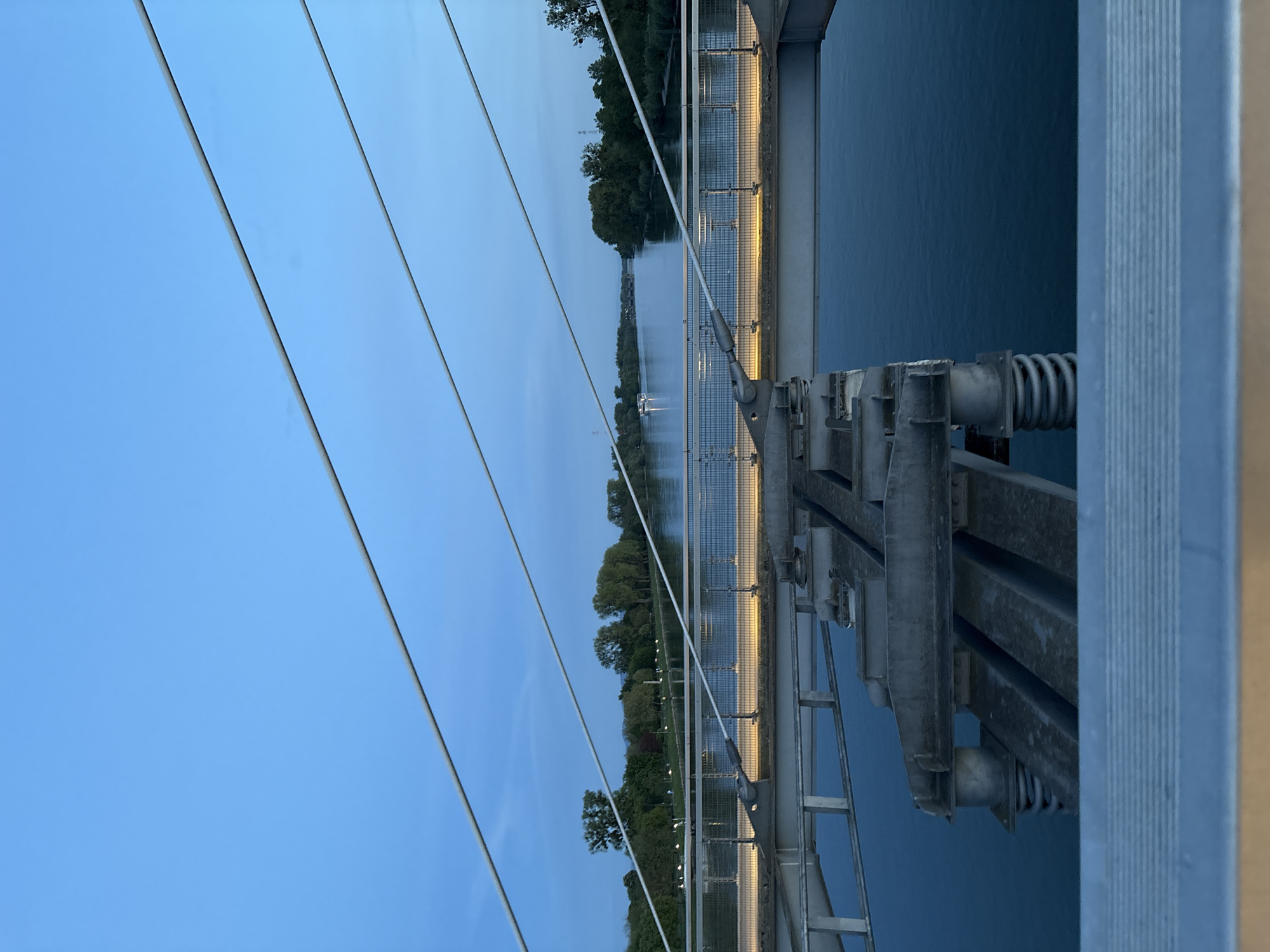The image captures a serene twilight scene from a modern bridge in Strasbourg, France, looking along its length over a calm body of water. In the foreground, several metallic suspension cables or guy-wires stretch diagonally across a clear, deep blue sky, creating a strong leading line. To the right, robust metal components of the bridge's structure, including railings and support beams, are visible, suggesting a contemporary architectural design.
Below the bridge, the tranquil water surface reflects the dimming sky. Along the opposite side of the bridge deck or a parallel walkway, numerous artificial lights are illuminated, casting a warm, yellow glow that reflects softly on the water. Further in the distance, a dense treeline borders the banks of the waterway. A small boat, identifiable by a solitary light, navigates the water in the middle distance. The lighting conditions indicate dusk or early evening, as the sky is still light blue but artificial lights are active. The scene conveys a peaceful and quiet atmosphere. There is no visible text in the image.