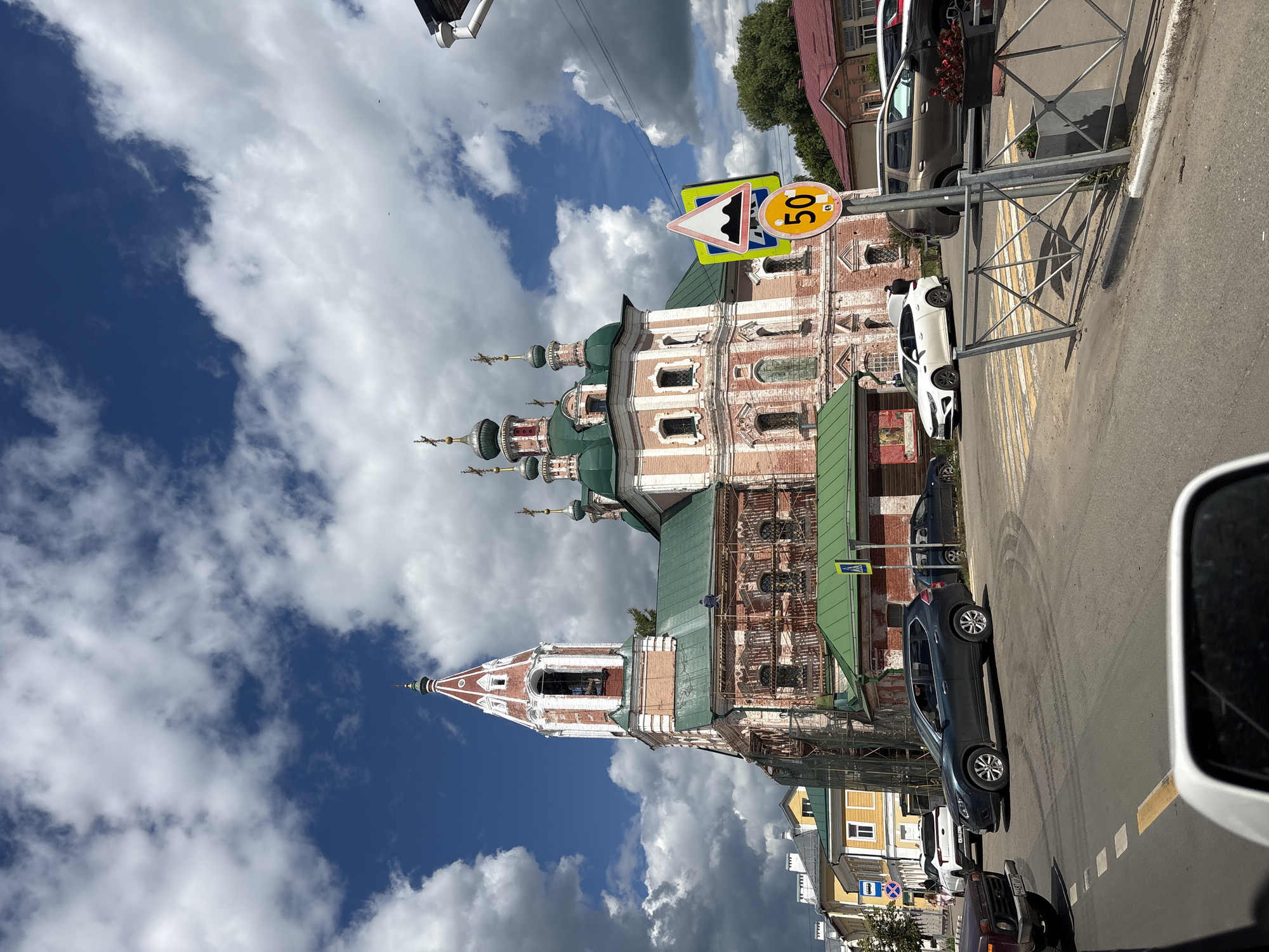 The image shows a street scene in Pereslavl-Zalessky, Russia, on a cloudy day. In the foreground, a dark-colored car is parked on the right side of the road. Further down the street, a white car is parked, and a person can be seen walking nearby. On the left side of the road, a historic brick building with green domes and white ornamentation, likely a church, is prominently featured. The building appears to be undergoing some restoration work, with scaffolding visible in parts of it. A speed limit sign displaying "50" is prominently displayed on a post, along with a triangular warning sign indicating a bumpy road. The sky is filled with dramatic, white and grey clouds against a deep blue background. The overall impression is that of a typical day in a Russian town, with a mix of modern vehicles and ancient architecture. The lighting suggests it is daytime, likely late morning or early afternoon.
