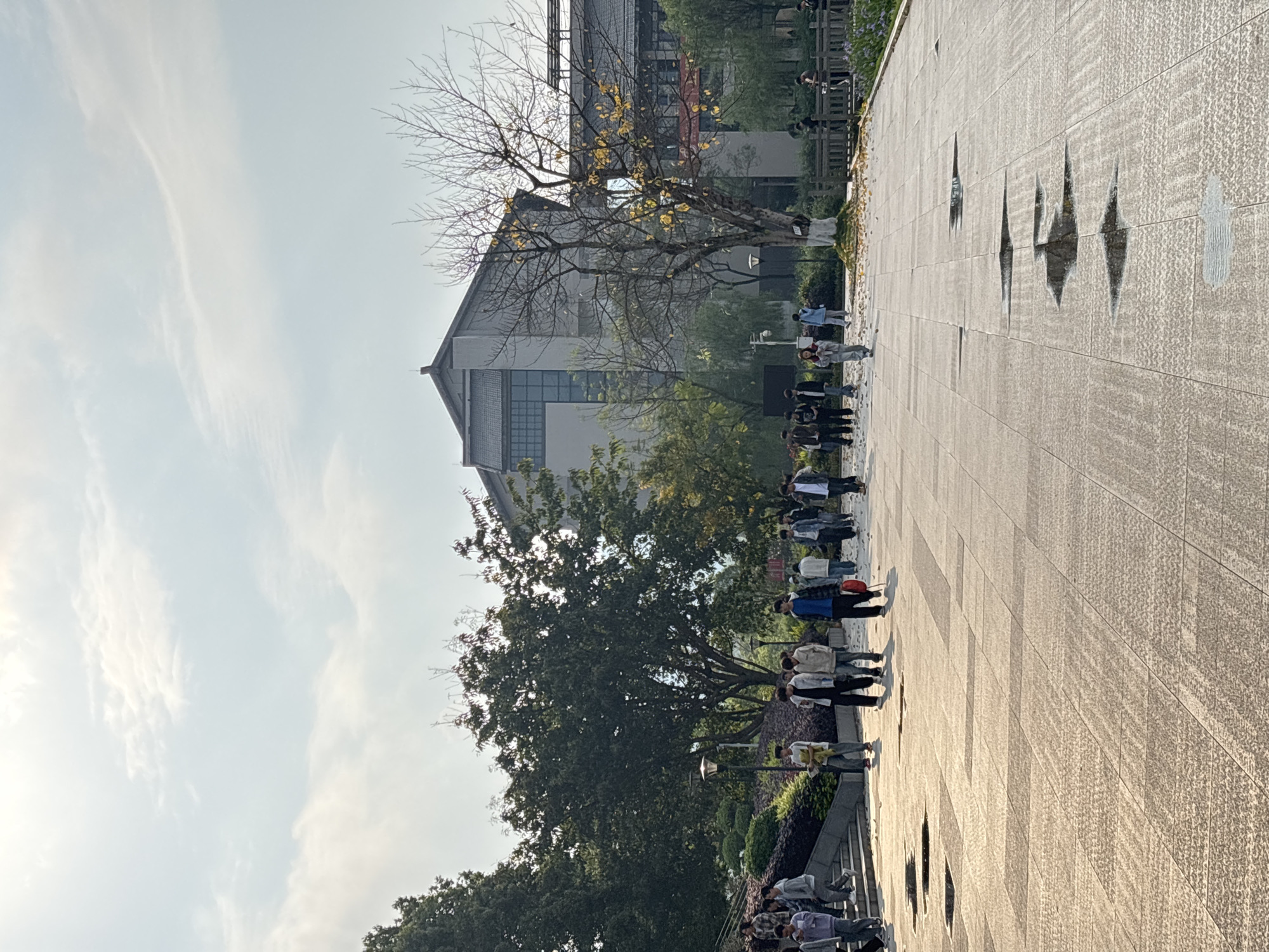 The image shows a group of people standing on a paved walkway in Jiangjin, China. The walkway is made of light-colored square stones with a textured surface. A building with a traditional Chinese roofline is visible in the background. Trees are interspersed along the walkway, some with sparse leaves. The sky is pale with light clouds. The image is taken from a ground-level perspective and appears to be rotated slightly.