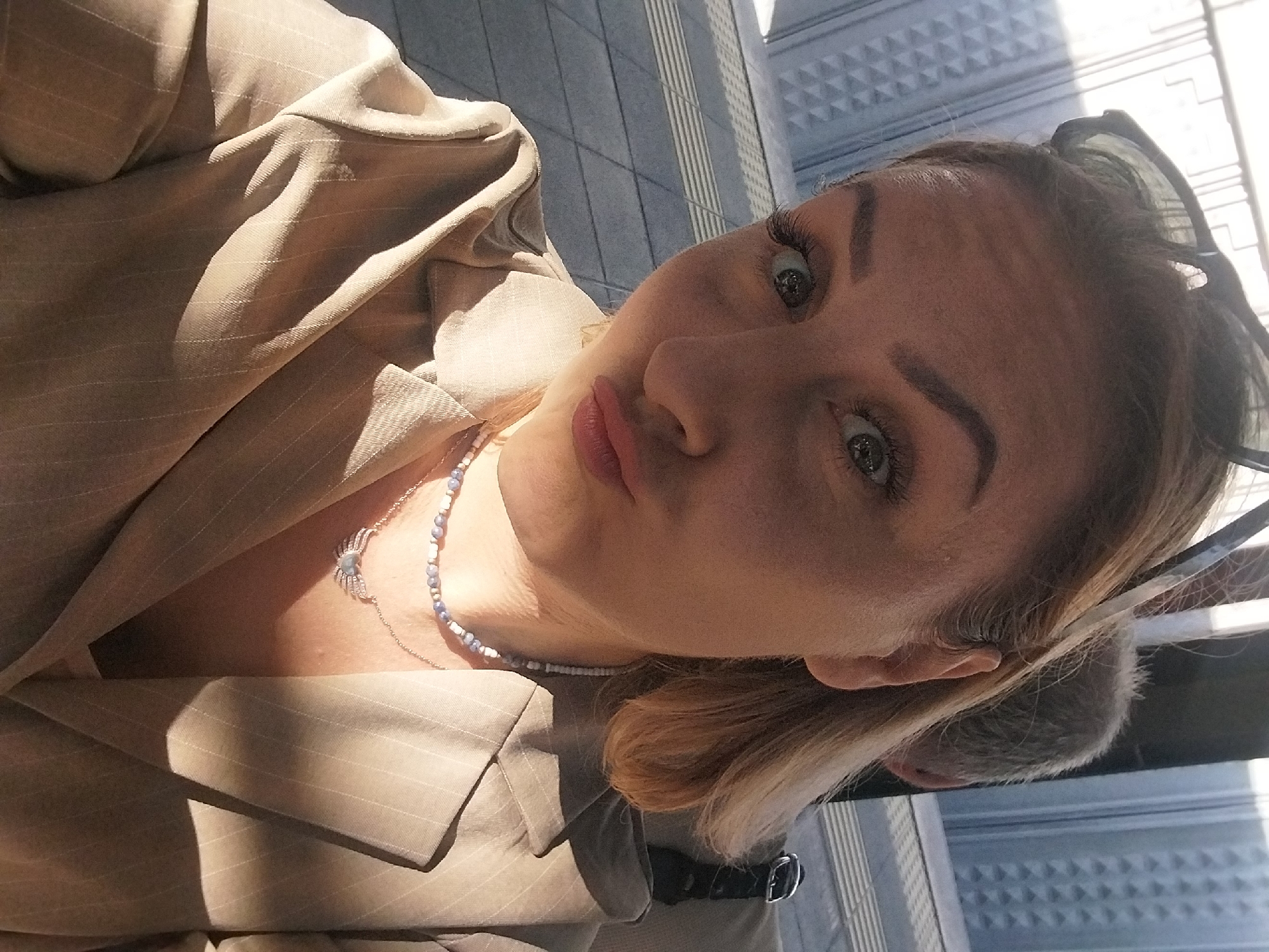 A woman is taking a selfie while sitting next to a person with grey hair. The woman has blonde hair, blue eyes, and is wearing a light-colored jacket, a beaded necklace, and sunglasses on her head. The background appears to be an urban setting with a building featuring a geometric pattern. The image is taken indoors, possibly on public transportation or in a cafe with large windows. The woman is making a duck face. The location is identified as Bakırköy, Turkey.
