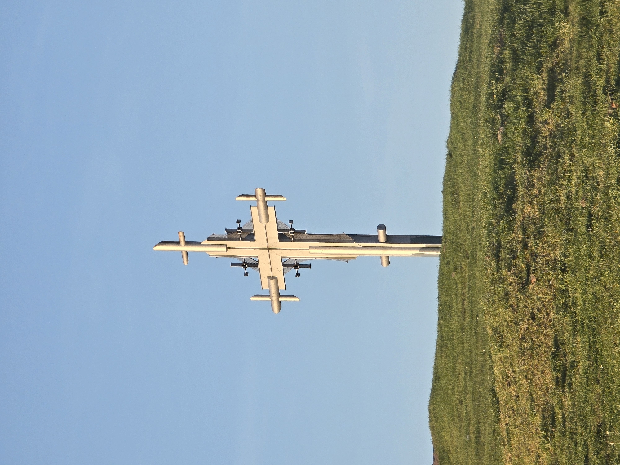 The image captures a striking metallic monument or sculpture positioned outdoors against a clear blue sky and a grassy landscape. The central subject is a tall, elaborate structure made of polished, light-colored metal beams. It features a prominent vertical column supporting a wide horizontal crossbeam. Several smaller, rounded projections extend from both the main horizontal beam and other parts of the structure, giving it a somewhat industrial or abstract, cross-like appearance. Intricate details, possibly lighting elements or sensors, are visible beneath the primary horizontal beam.
To the right of the monument, a vibrant green lawn covers a gently sloping area, suggesting a park or open field setting. The expansive sky, a uniform and clear light blue, dominates the left and upper background, providing a serene backdrop for the metallic edifice.
The soft, natural lighting suggests either the late afternoon or early morning, casting subtle shadows and highlighting the metallic surfaces. No specific activity is occurring; the scene depicts a static, monumental installation. There is no visible text within the image. Based on the provided context, this scene is located in Burgas, Bulgaria.
