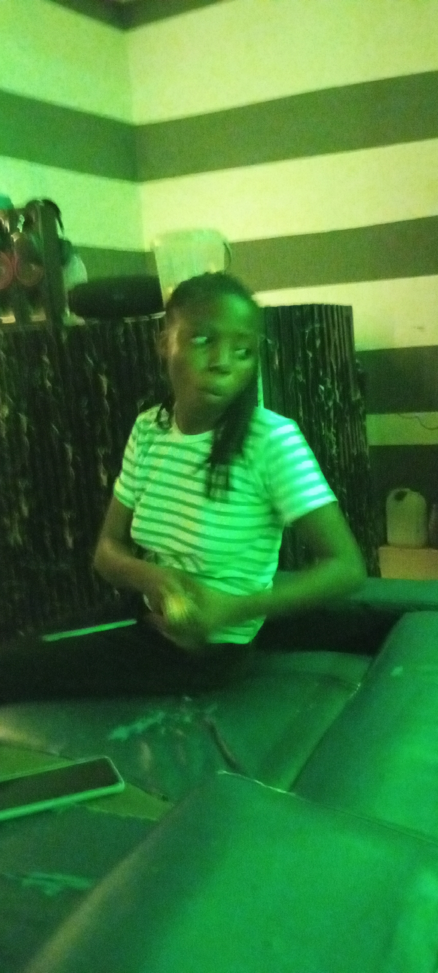 In Adankolo, Nigeria, a young woman with dark hair, wearing a green and white horizontally striped t-shirt and dark pants, is seated. She is holding a small yellow object in her hands, which are positioned in front of her chest. Her gaze is directed towards her left. The environment is dimly lit with a strong green hue. In the background, striped walls are visible, with dark, vertical panels interspersed. To the left, there are hints of gym equipment, including circular weights. A dark couch or seating area is in the foreground, with a dark rectangular object, possibly a phone, placed on its surface. The overall impression is an indoor setting, possibly a gym or lounge area.
