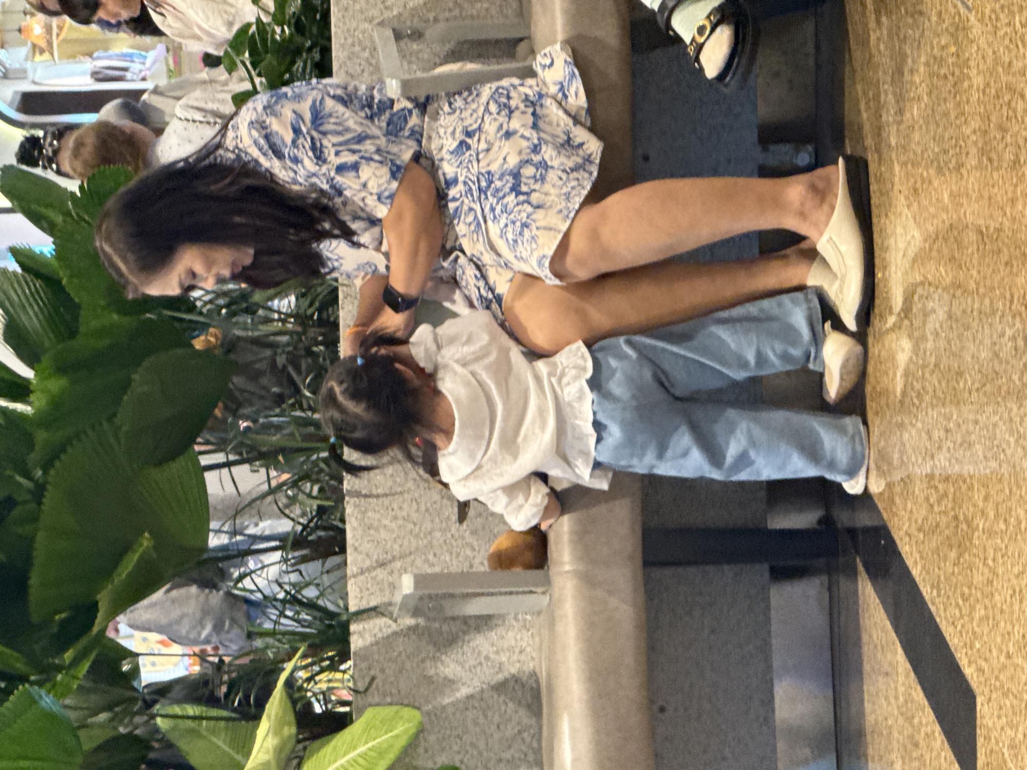 The image captures a mother and daughter in a public indoor space with lush greenery. The mother, wearing a blue and white patterned dress, is seated on a beige bench, attending to her daughter. The daughter, dressed in a white ruffled top and light blue pants, stands facing away from the camera, towards the mother. She has her hair in pigtails. In the background, other people and potted plants are visible. The floor is a reflective brown material. The scene is likely a waiting area or a shopping mall in Bắc Cam Ranh, Vietnam.