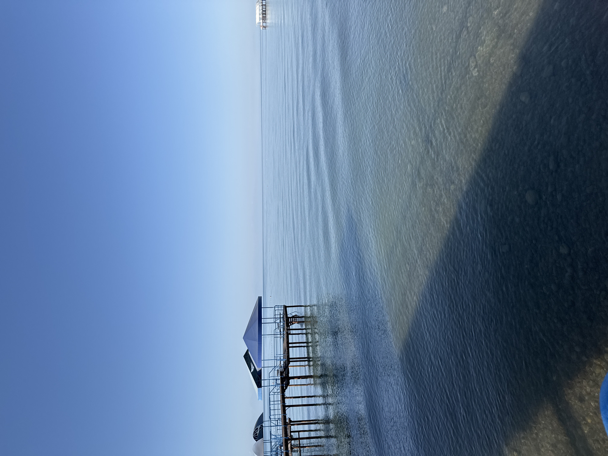 The image shows a tranquil scene of a pier extending into a calm body of water under a clear blue sky. The pier, with its wooden planks and blue railings, features covered seating areas. The water is shallow near the shore, revealing the pebbly bottom, and becomes deeper further out. In the distance, a small structure on stilts is visible on the water. The overall environment suggests a serene waterfront location, likely a beach or a lakeside resort, possibly in Sary-Oy, Kyrgyzstan, known for its location on the shores of Issyk-Kul Lake.