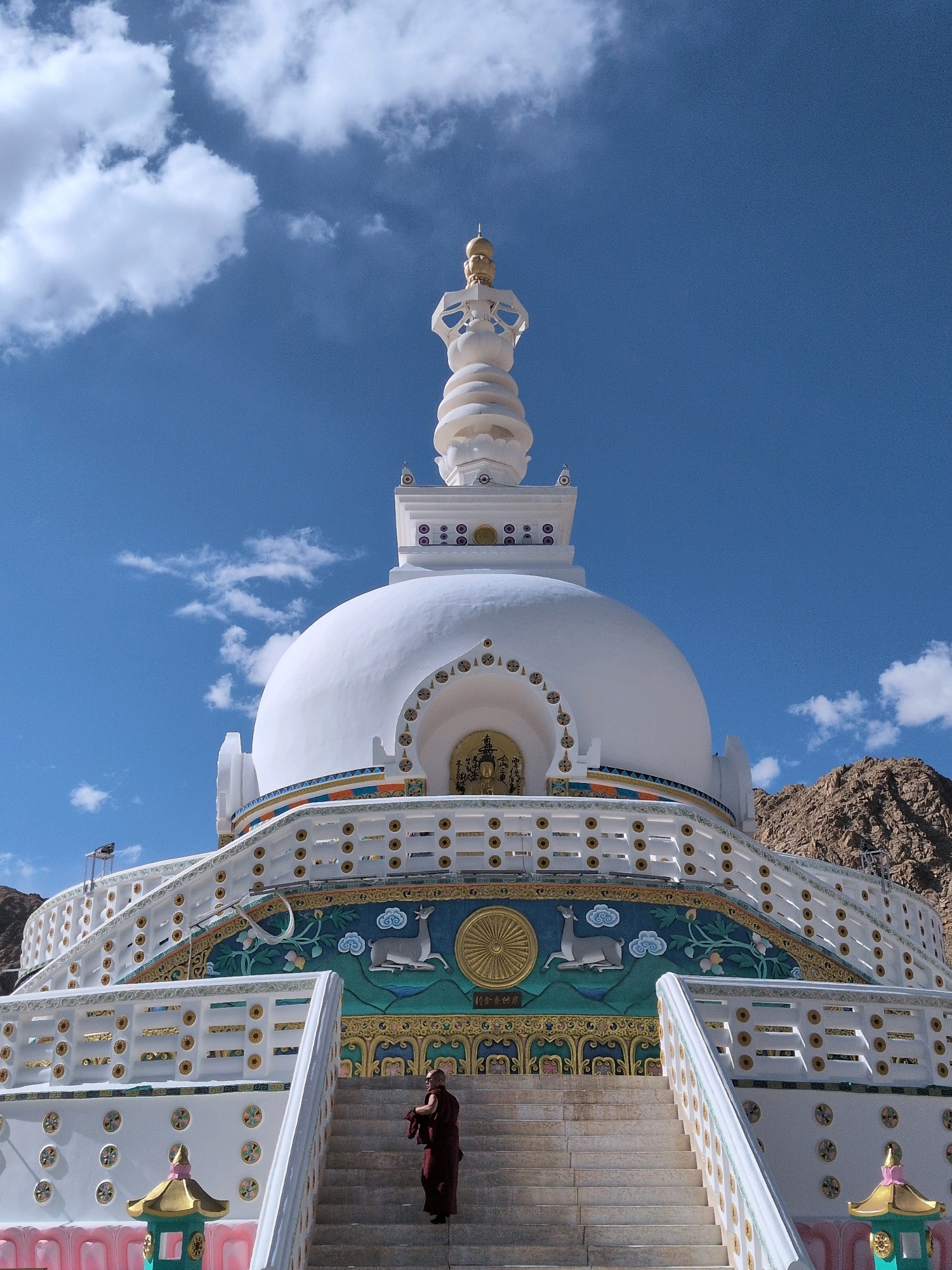 The image shows the Shanti Stupa in Leh, India, a Buddhist white-domed stupa situated on a hilltop. The stupa, with its golden spire, is a prominent landmark against a clear blue sky dotted with white clouds. The structure features intricate decorative bands with colorful murals depicting Buddhist motifs, including deer and a Dharma wheel. A monk dressed in maroon robes is ascending a wide set of stairs leading up to the stupa. The foreground shows the lower portion of the stupa's tiered base and some decorative elements, while the background features the rugged, arid mountains characteristic of the region. The overall scene conveys a sense of peace and spiritual significance.
