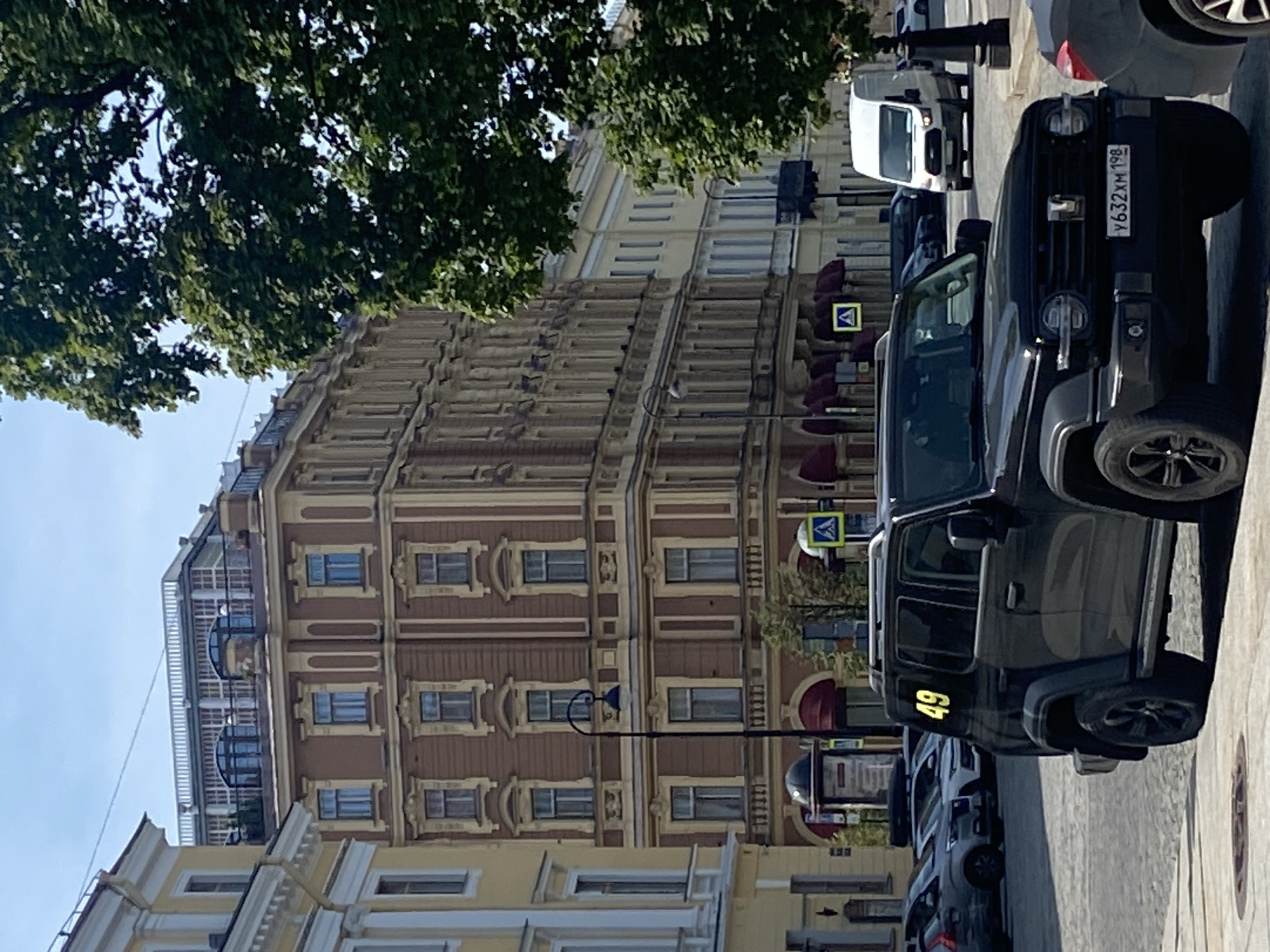 The image captures a street scene in Saint Petersburg, Russia, characterized by historic architecture and vehicular traffic. In the foreground, a black SUV is parked, displaying the number "49" in yellow on its side and a license plate reading "Y632XM 198". Behind the SUV, a white van is visible, and several other cars are parked or driving on the cobblestone street.
The background features ornate buildings with multiple stories, exhibiting classic European architectural styles. The building on the left is painted a light yellow, while the larger building in the center is a rich brown with yellow accents. Several windows, awnings, and decorative elements are discernible on these structures. A lamppost stands prominently in front of the brown building. Lush green trees line the street, adding a touch of nature to the urban environment. The sky is clear and blue, indicating a sunny day. The overall impression is of a bustling city street with a blend of historical charm and modern life.