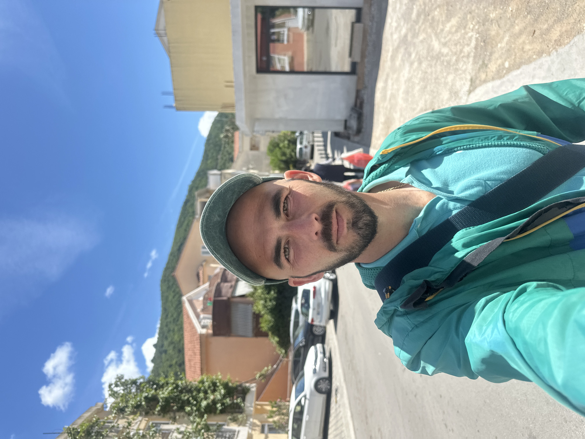 The image is a selfie taken outdoors on a sunny day. The person in the photo is a fair-skinned man with short brown hair, a beard, and brown eyes. He is wearing a blue baseball cap, a light blue shirt, and a green jacket with yellow trim. He has a dark blue strap across his chest.
The background shows a street with buildings and parked cars. There are trees and a mountain in the distance. The buildings appear to be residential and commercial properties.
Based on the architecture and landscape, the location is likely in Budva, Montenegro. The photograph captures a casual moment, possibly during a walk or outing.