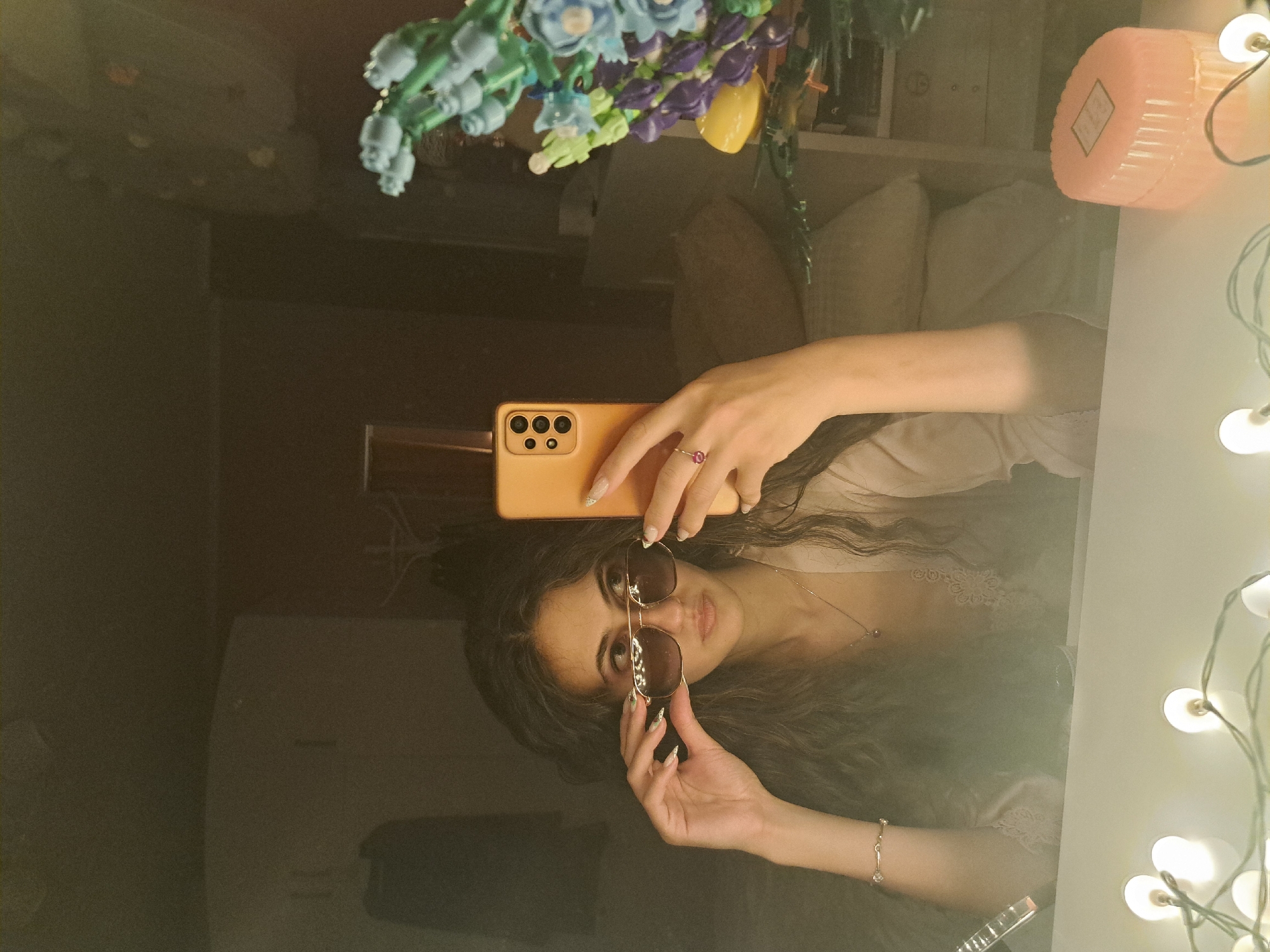 This image is a mirror selfie featuring a young woman captured indoors in Negovan, Bulgaria. She is seen from the chest up, holding an orange-cased smartphone in her left hand to take the photo, and adjusting a pair of aviator-style sunglasses with her right hand. She has long, dark, wavy hair and looks directly at the viewer through the sunglasses. Her left hand wears a ring with a prominent pink stone, and her right wrist has a silver bracelet. Her fingernails are adorned with an intricate white and green design. She is wearing a light-colored top, possibly with lace trim, and a delicate necklace.
The reflection reveals elements of the room. Above the woman's head, an arrangement of artificial flowers in shades of blue, green, purple, and yellow is visible. To the right, a string of warm white decorative lights illuminates the scene, alongside a pink, ribbed cylindrical container. A label on the container is present but the text is illegible. The background of the reflection includes white pillows and what appears to be a white safe or cabinet. The overall lighting is warm and soft, suggesting an indoor environment, possibly in the evening or a dimly lit space. The reflective surface itself is dark and polished.