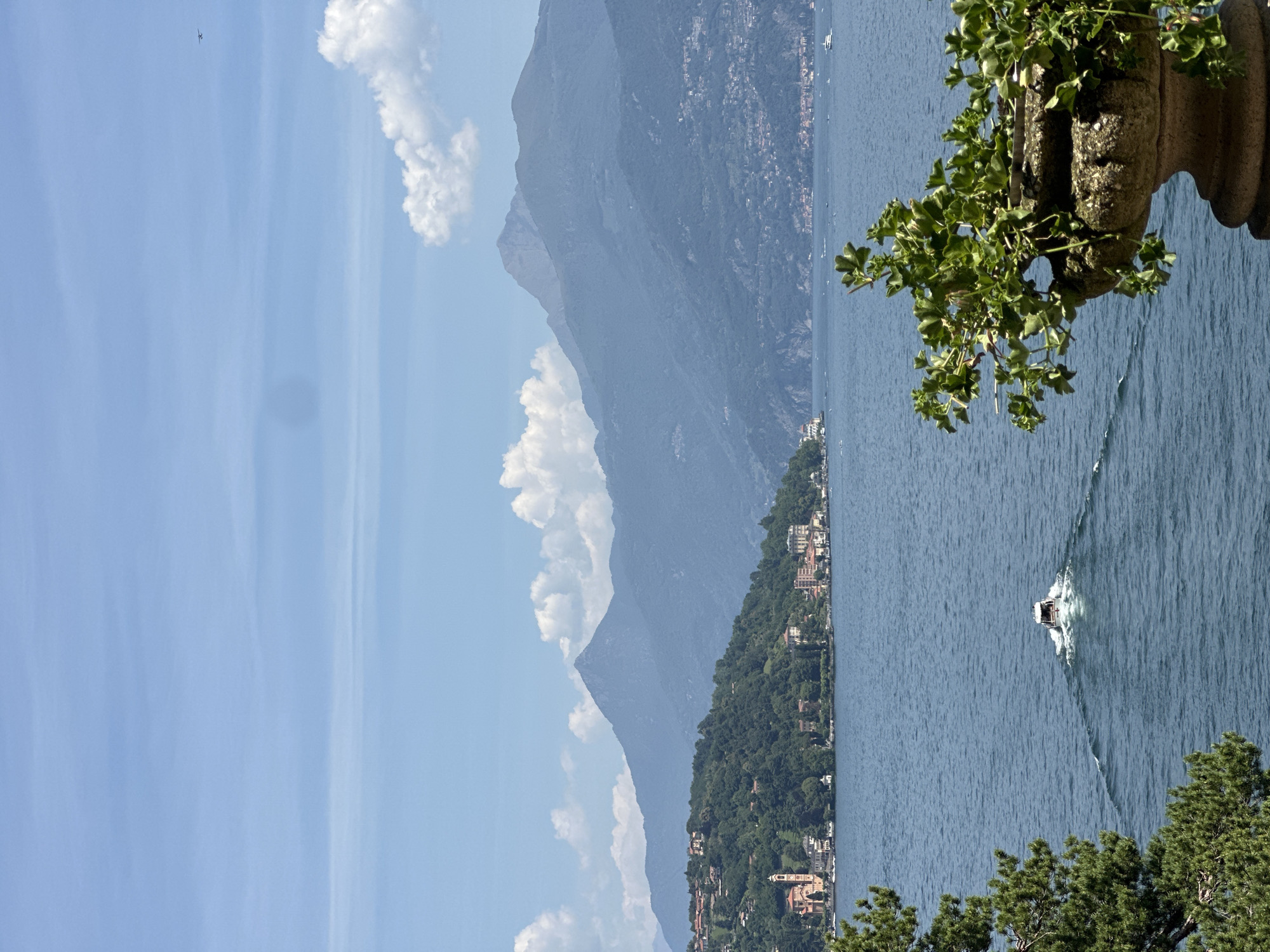 This is a landscape photograph of Lake Como in Tremezzina, Italy. The foreground features a stone planter with green foliage. In the mid-ground, a white boat is moving across the blue water, leaving a wake behind it. The background consists of a lush green hillside dotted with buildings, a prominent church tower, and a chain of misty blue mountains under a hazy sky with scattered white clouds. A small airplane is visible in the upper right corner of the sky.
