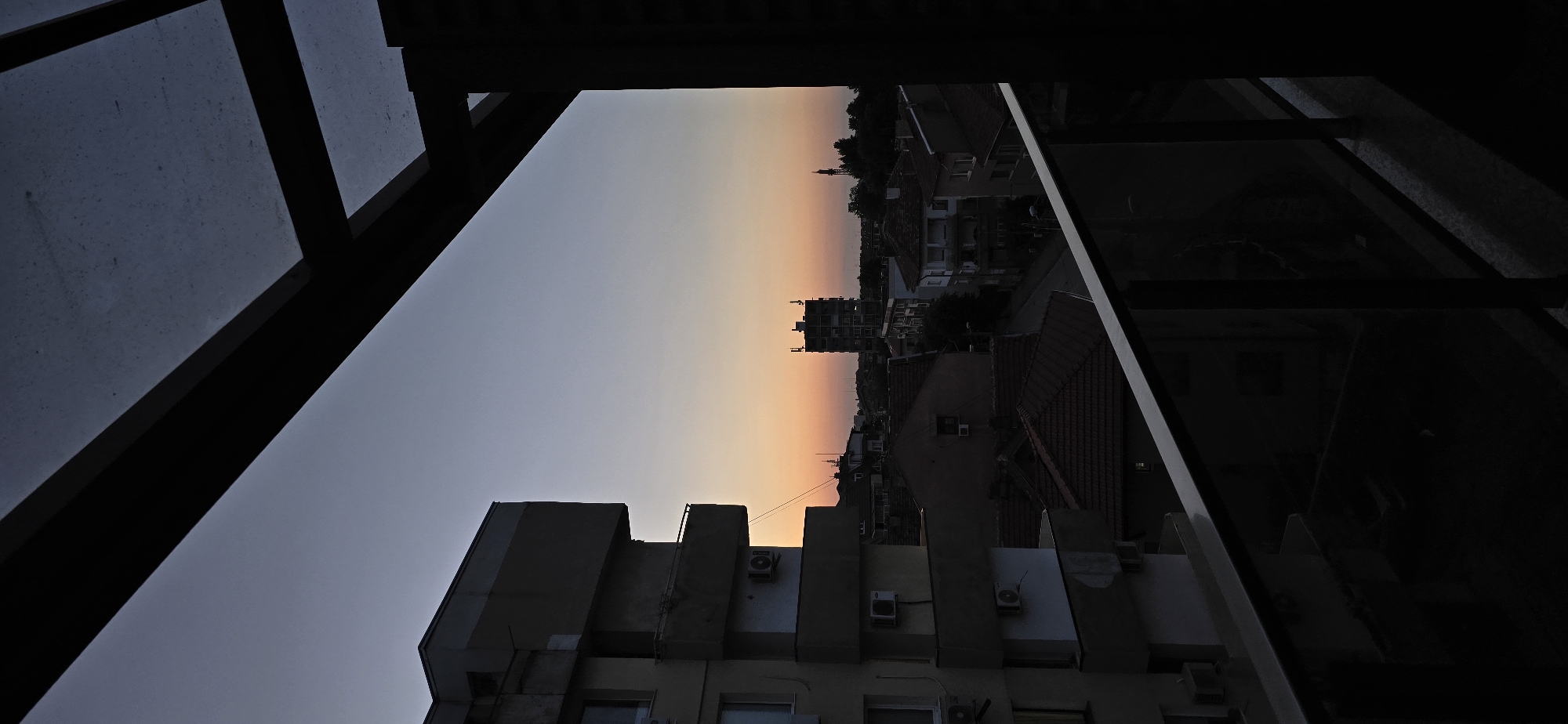 A high-angle urban landscape captured from a balcony at either dusk or dawn. The sky displays a gradient, transitioning from a dark blue-grey at the zenith to vibrant orange and pink hues along the horizon.
In the upper right, a dark structural overhang with visible beams and a translucent, textured roof panel frames the top of the image. The immediate foreground on the left features a portion of a multi-story building facade, heavily shadowed, displaying several air conditioning units and balconies. Across the bottom, a dark metal railing with clear panels delineates the balcony edge.
The midground reveals a dense urban environment. To the left, a multi-story residential building, similar to the perspective structure, stands predominantly in shadow, with visible balconies and exterior air conditioning units. Further into the scene, a cluster of residential buildings with various roof styles (some tiled, some flat) occupies the central and right portions, mostly appearing as dark silhouettes. A street is discernible, winding through these buildings. In the central distance, a tall, rectangular building with multiple floors and roof-mounted structures (possibly antennas or dishes) is silhouetted against the colorful sky. To its right, a slender, pointed tower, resembling either a minaret or a communication tower, is also silhouetted against the horizon, partially obscured by trees.
The entire scene is characterized by the low, ambient light of either sunrise or sunset, creating strong contrasts and deep shadows, particularly in the foreground and on the city structures. No individuals are visible. The setting is an urban environment within the city of Unknown, Unknown.
