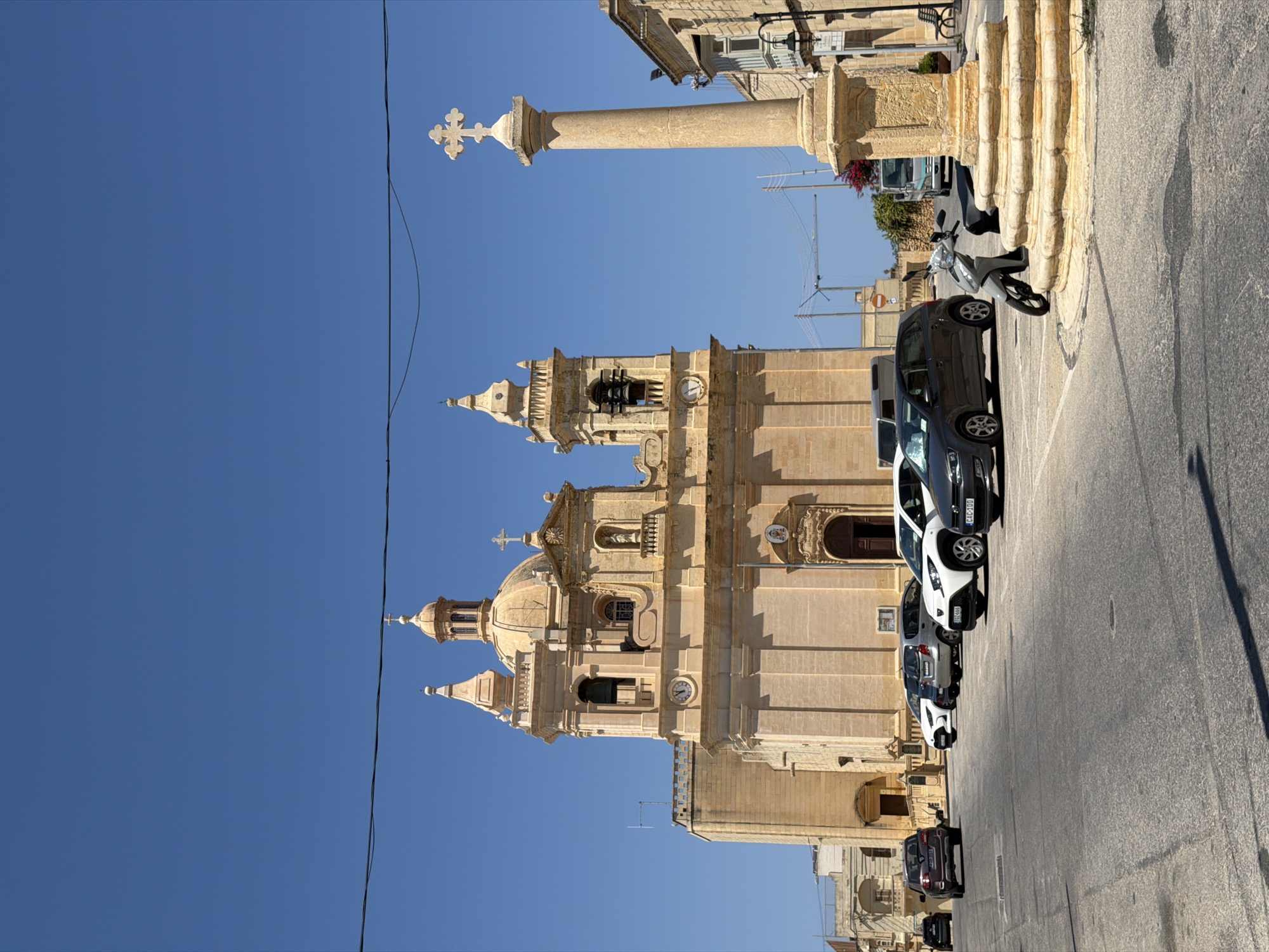 In the city of Żebbuġ, Malta, a clear, bright day unfolds in a town square. The focal point is a large, pale stone church with a dome and two bell towers, its facade adorned with architectural details. In front of the church, a row of parked cars, including a dark SUV, a white hatchback, and a grey hatchback, occupy the paved square. A stone monument, a tall, weathered column topped with a cross, stands to the right of the church. A light-colored scooter is parked near the base of the monument, its kickstand down. The sky above is a vibrant, cloudless blue, and a black wire stretches across the top of the frame. Beyond the immediate square, hints of other buildings, construction cranes, and distant foliage add to the urban landscape.