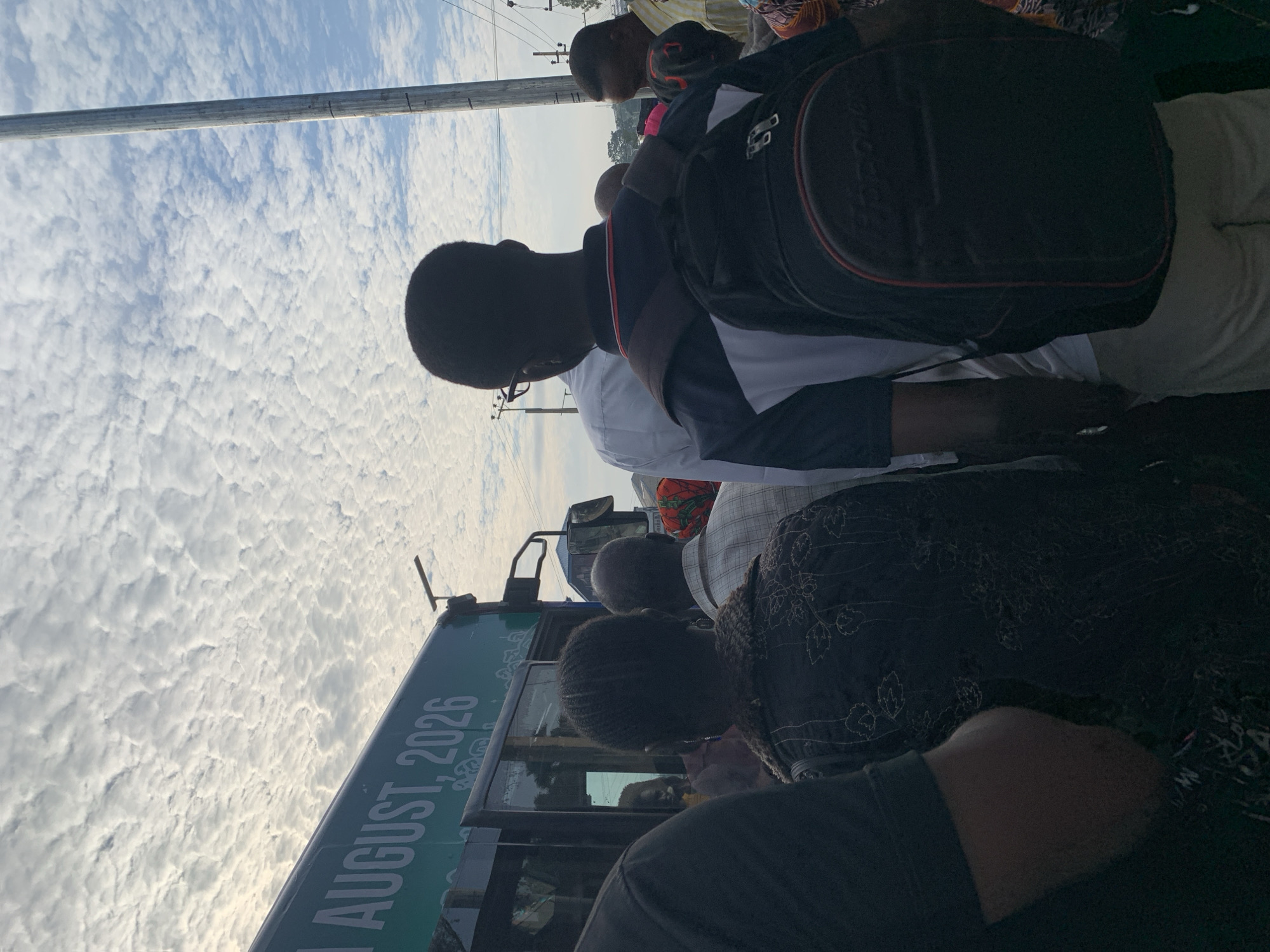 The image shows a group of people standing in what appears to be a queue, possibly waiting to board a bus. The bus is green and has the text "1 AUGUST, 2026" visible on its side. The sky is filled with scattered, puffy white clouds, suggesting it might be daytime. In the background, a metal pole, possibly a utility pole, is visible, with some wires or cables attached. The people are mostly seen from behind, with one person in the foreground wearing a black backpack. The overall scene seems to be set in an outdoor urban or semi-urban environment. The location is stated to be Rafin Par, Nigeria.
