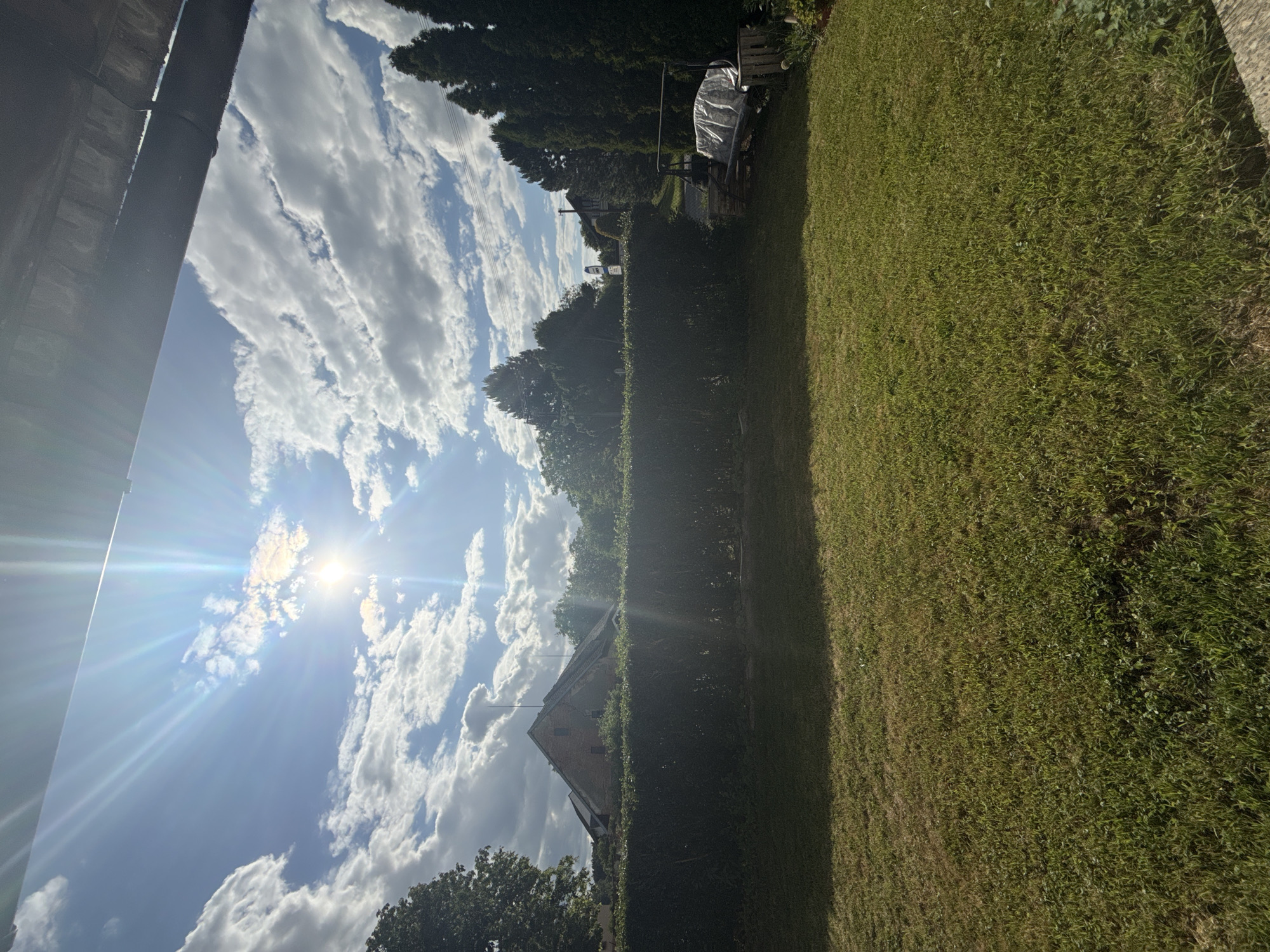An outdoor scene captured under a bright, partly cloudy sky, featuring a sunlit residential or rural backyard. The sun, high in the upper-middle left, emits strong rays and significant lens flare. The sky is a light blue, adorned with numerous white cumulus clouds. The very top edge of the image shows the underside of a dark, indeterminate overhead structure, possibly a roof eave or awning.
The immediate foreground is a verdant, recently mown grass lawn, exhibiting areas of both lush green and drier, lighter patches. A dense, dark green trimmed hedge forms a horizontal barrier across the midground. Behind the left section of the hedge, a large, dark-leafed deciduous tree is visible. Further back and to the left, a stone or brick building with a steeply pitched, dark roof and small windows stands; several antenna-like structures project from its roofline. Additional trees are visible beyond this building.
Behind the right section of the hedge, a row of tall, slender evergreen trees with dark foliage forms a backdrop. To the right of the hedge, a metal A-frame swing structure is present, its seat covered by a light-colored tarp or sheet. Adjacent to the swing, a light-toned wooden crate or planter is placed. Beyond the hedge and trees on the right, overhead power lines traverse the upper background, and glimpses of a path or road and distant structures are discernible. No individuals or animals are present in the frame. The location is in the city of Unknown, Unknown.