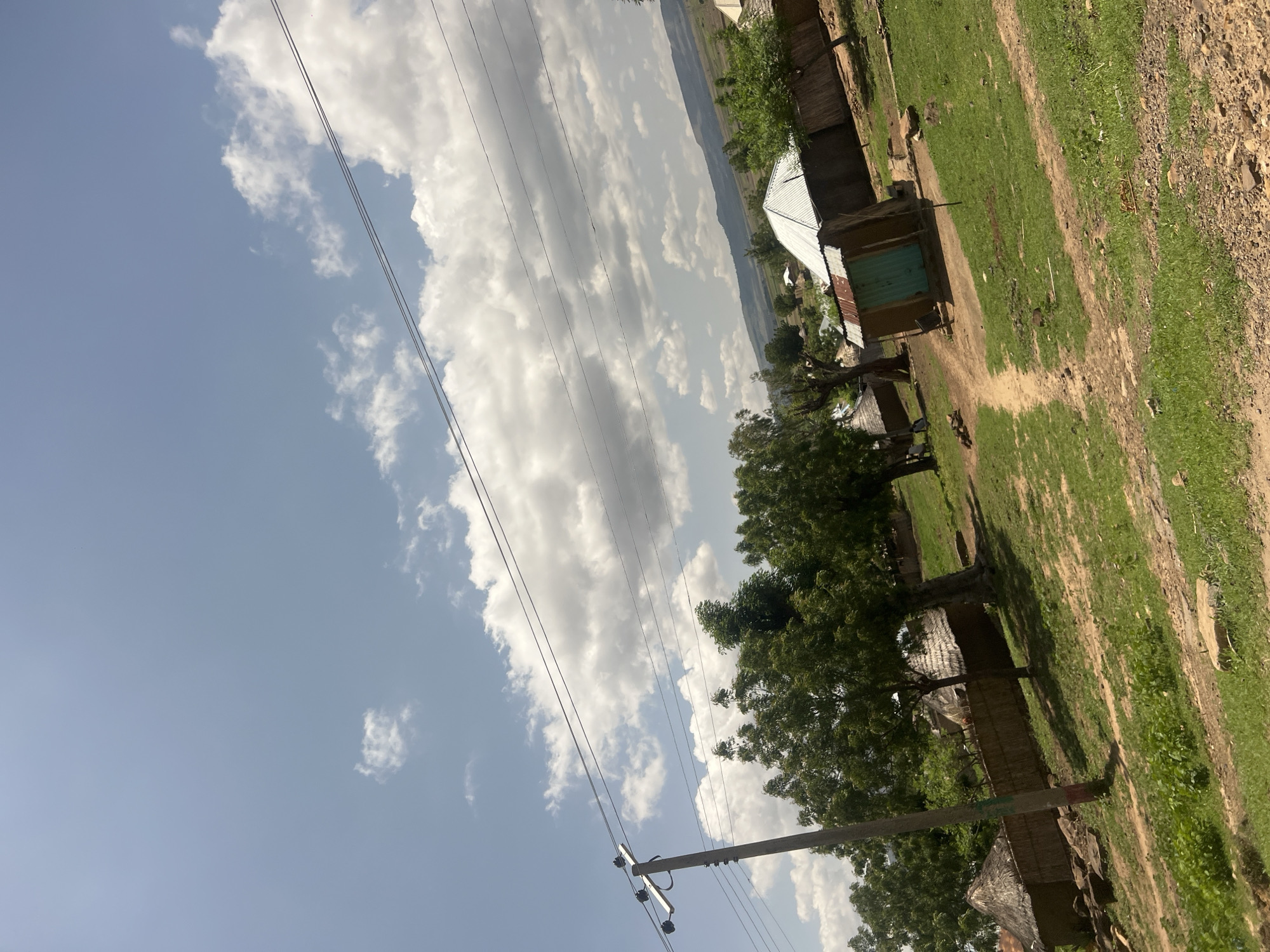 A rural landscape in Chum, Nigeria, is depicted under a bright, partly cloudy sky. The upper half of the image features a clear blue sky with scattered white cumulus clouds. A wooden utility pole with multiple overhead power lines extends diagonally from the lower-left, with the lines traversing horizontally across the mid-upper section of the frame. Below the power lines, numerous green trees with dense foliage are prominent, particularly in the left and central areas. The ground is a mix of green grass, lighter dirt paths, and exposed soil patches with scattered rocks.
Several structures are visible across the scene. On the far left, partially obscured by the utility pole, are two round huts with thatched roofs and walls constructed from natural, possibly woven, materials, situated behind a low fence. Towards the center-right, a rectangular building with a distinct silver corrugated metal roof and solid, possibly concrete or mud, walls stands out. Adjacent to this structure is a smaller, rustic building with a turquoise-colored door and a corrugated metal roof. Other less distinct structures and fences are present further in the background. In the distant background, faint outlines of rolling hills or mountains are visible under a hazy sky. No specific individuals or discernible human actions are clearly present within the frame.