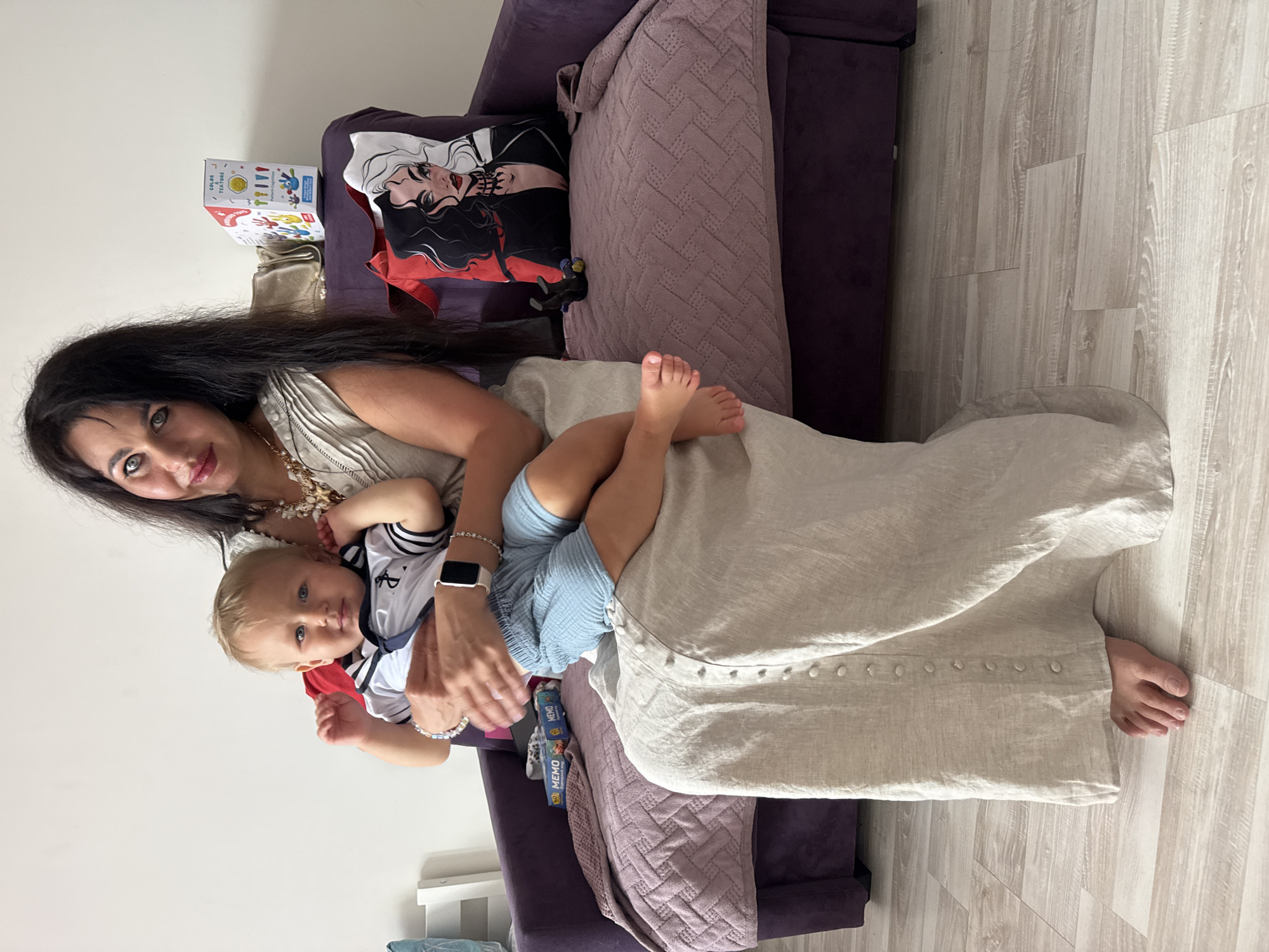 In a living room setting, a woman with long dark hair is holding a baby. The woman is wearing a beige sleeveless top and a pearl necklace, with a smartwatch on her left wrist. The baby is dressed in a sailor-style outfit with blue and white stripes, red accents, and a white collar. The baby has blond hair and blue eyes and is looking at the camera. The woman is smiling, and the baby has a neutral expression.
Behind them, a purple sofa is visible with several cushions. One cushion features a graphic print of two women with long dark hair. A textured pink blanket is draped over the sofa. On the floor next to the sofa, a child's game box with colorful graphics is visible, with the word "MEMO" printed multiple times. The flooring is light-colored wood planks. The scene appears to be indoors, possibly during the day, given the ambient lighting. The overall mood seems relaxed and intimate. The provided location context is Sochi, Russia.