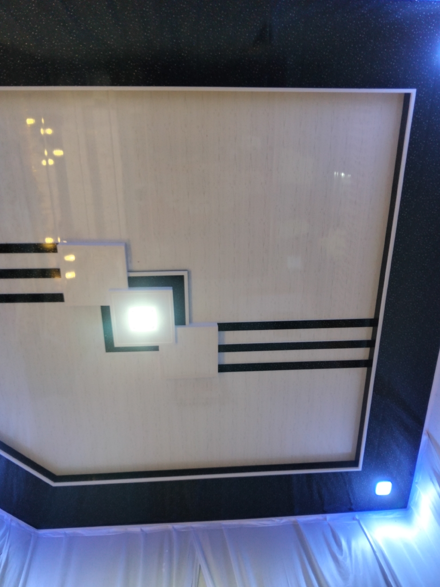 The image shows a modern ceiling design with recessed lighting and a dark, speckled border. The central part of the ceiling is a light-colored rectangular panel with a textured surface. Geometric shapes in white and black are integrated into the design, with thin black stripes accentuating the central panel. A bright square light fixture is centrally located within one of the geometric elements. The ceiling is illuminated by a bright, white light from this fixture and also by subtle blue lighting along the lower edges, near the curtains. The image is taken from a low angle, looking upwards, and the lower portion of the frame shows draped white curtains, likely part of an interior space such as a hall or room. There are no people visible in the image. The location is described as Cottrueng, Indonesia.