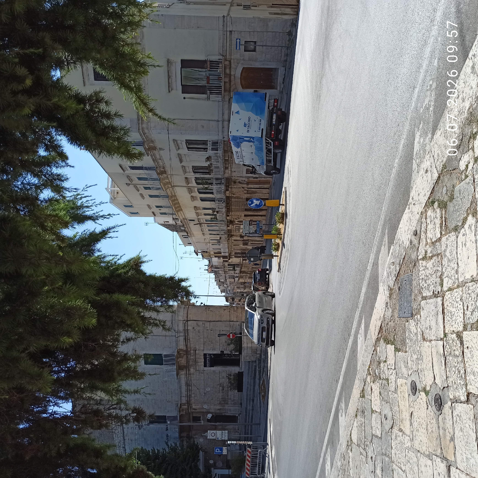 In Ruvo di Puglia, Italy, a street scene unfolds under a clear blue sky. The foreground features a cobblestone sidewalk with two circular metal manhole covers and one rectangular metal grate. A grey asphalt road with white lane markings dominates the midground, curving gently to the left. A silver minivan is turning left onto the road from the foreground. Further down the road, a dark SUV is traveling in the same direction as the minivan. On the right side of the road, a white truck with a blue and white geometric design on its side is parked. The truck has "DI TACCHIO" written on its side.
The background is lined with old buildings, characterized by their white stone facades, multiple balconies with white railings, and dark-shuttered windows. Some buildings have arched doorways and ornate architectural details. Trees with dark green foliage hang over the street from the left side of the frame, partially obscuring the view of the buildings. Traffic signs, including a blue sign with a white bird and directional arrows, are visible on the right side of the road. A traffic light and a "P" parking sign are present on the left side of the intersection. The image is time-stamped with "06.07.2026 09:57".