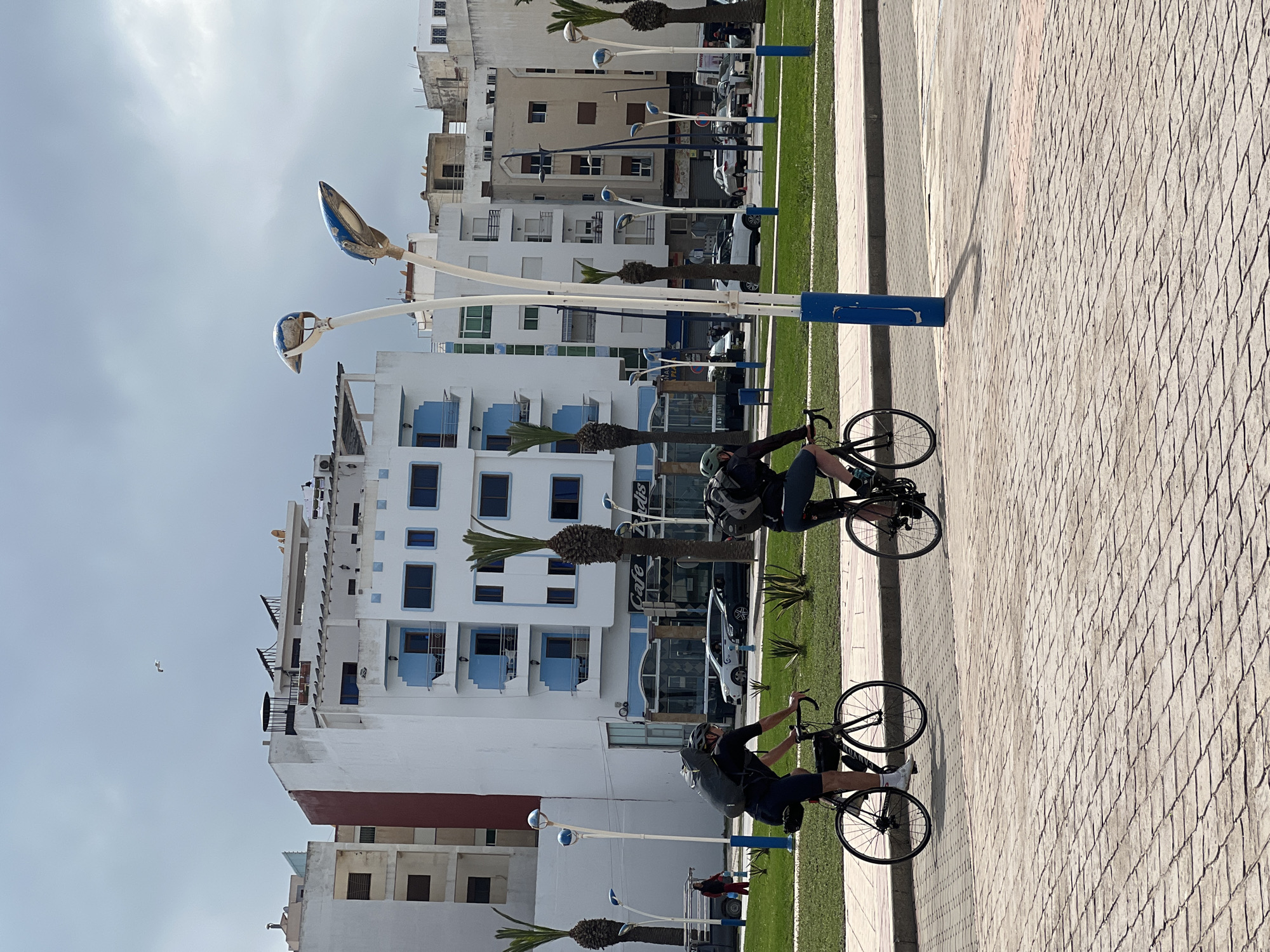 Two cyclists ride on a paved path next to a narrow strip of grass bordering a street. The buildings along the street are mostly white with blue accents and various architectural styles, including apartments and commercial spaces, such as a "Cafe Paris." Palm trees are planted at regular intervals along the street. Overhead lighting fixtures line the street and a gull flies in the sky. The image location is M'Diq ⵎⴹⵢⴰⵇ المضيق, Morocco.