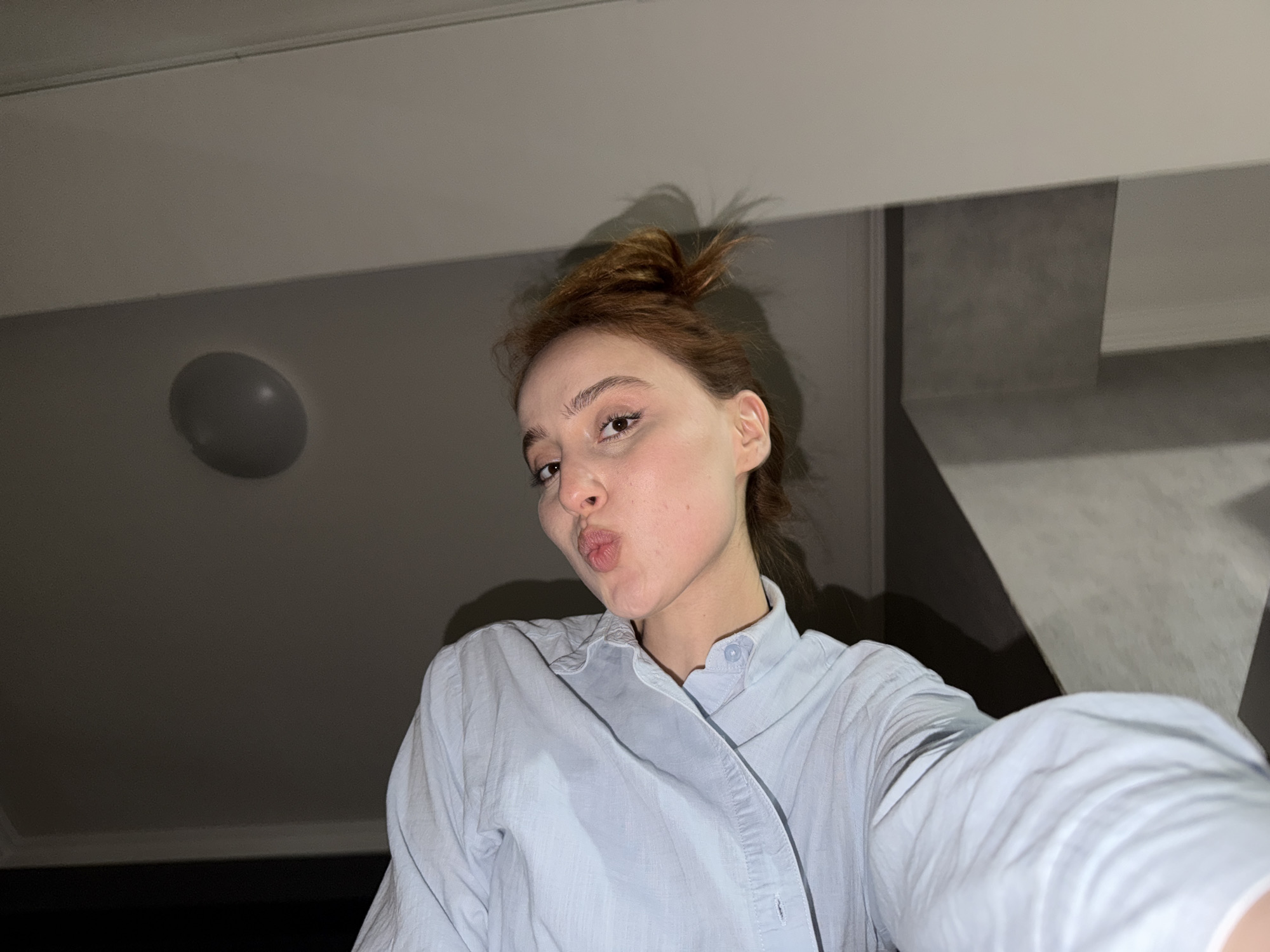 A young woman is positioned in the lower center of a medium, low-angle, close-up shot taken indoors. She is looking into the camera with pursed lips. Her hair is reddish-brown and is pulled back into a messy bun. She is wearing a light blue button-down shirt. The background reveals a minimally decorated room with a light fixture on the ceiling to the left and a vertical structural element to the right. The scene appears to be illuminated by artificial light, contributing to the even lighting across the surfaces. Given the style, the photo appears to be a selfie. The location details (Tbilisi, Georgia) are not discernible from the visual information provided.