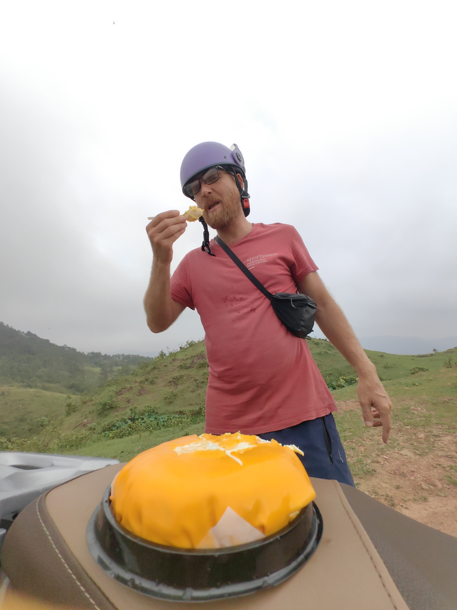 The image displays a man taking a bite of a bright yellow dessert, likely a cake, in a hilly outdoor setting. The man has a beard and wears glasses, a purple helmet with a small accessory mounted on top, a red short-sleeved t-shirt, blue shorts, and a black fanny pack worn cross-body. He holds a small wooden spoon to his mouth, which contains a portion of the dessert.
The partially eaten yellow cake, revealing white creamy layers, sits in a black plastic container in the foreground, resting on the brown, stitched seat of what appears to be a scooter or motorcycle. The man's red t-shirt has the word "sportswear" printed on it, along with smaller, less legible text below.
The background features a vast, lush green landscape with rolling hills and distant valleys, indicating a rural or mountainous area. The sky is uniformly overcast and light grey, suggesting a cloudy day. The ground beneath the man's feet is a mix of green vegetation and reddish-brown soil. The scene conveys a moment of enjoyment during a stop in a scenic, natural environment, consistent with locations like Cao Xanh Ward, Vietnam.